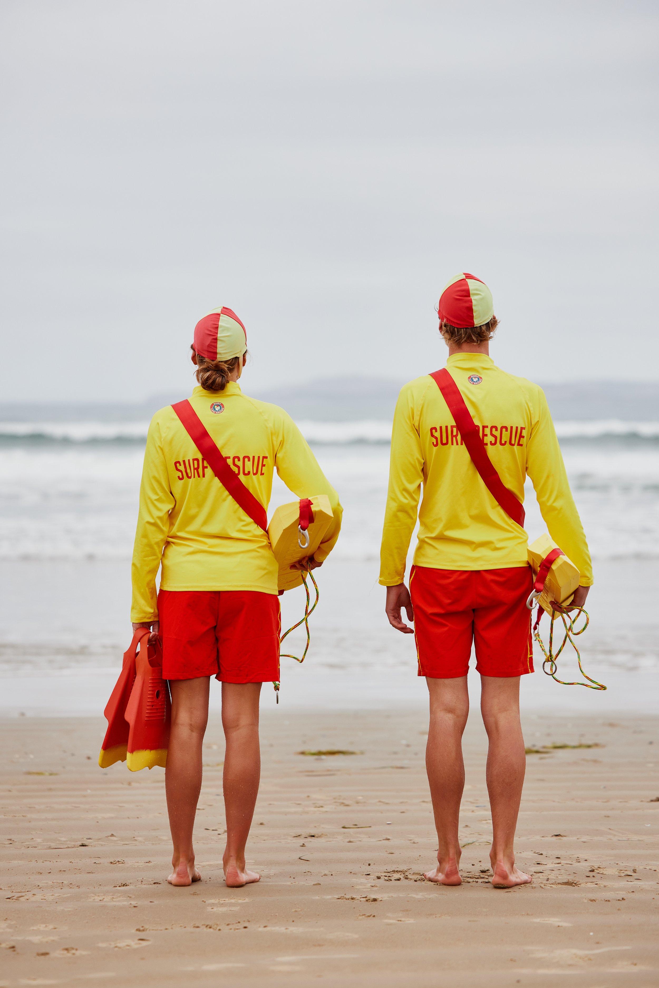 Two lifeguards in yellow and red uniforms and hats standing on the beach, facing the ocean, ready to perform water rescue.