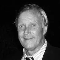 Black and white photograph of a man with light-colored hair, wearing a suit and tie, smiling at the camera.