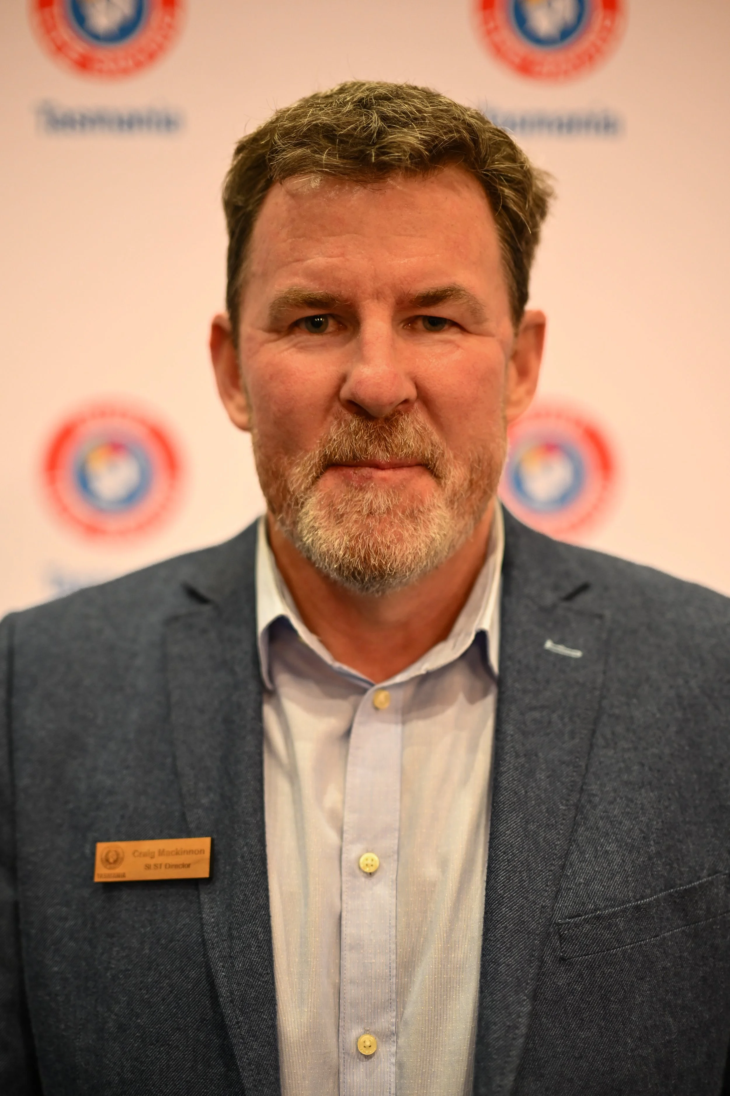 A middle-aged man with brown hair and a beard, wearing a gray suit jacket and a light-colored shirt, standing in front of a white wall with multiple logos and a name tag that reads 'Craig Mackinnon, SI ST Director'.