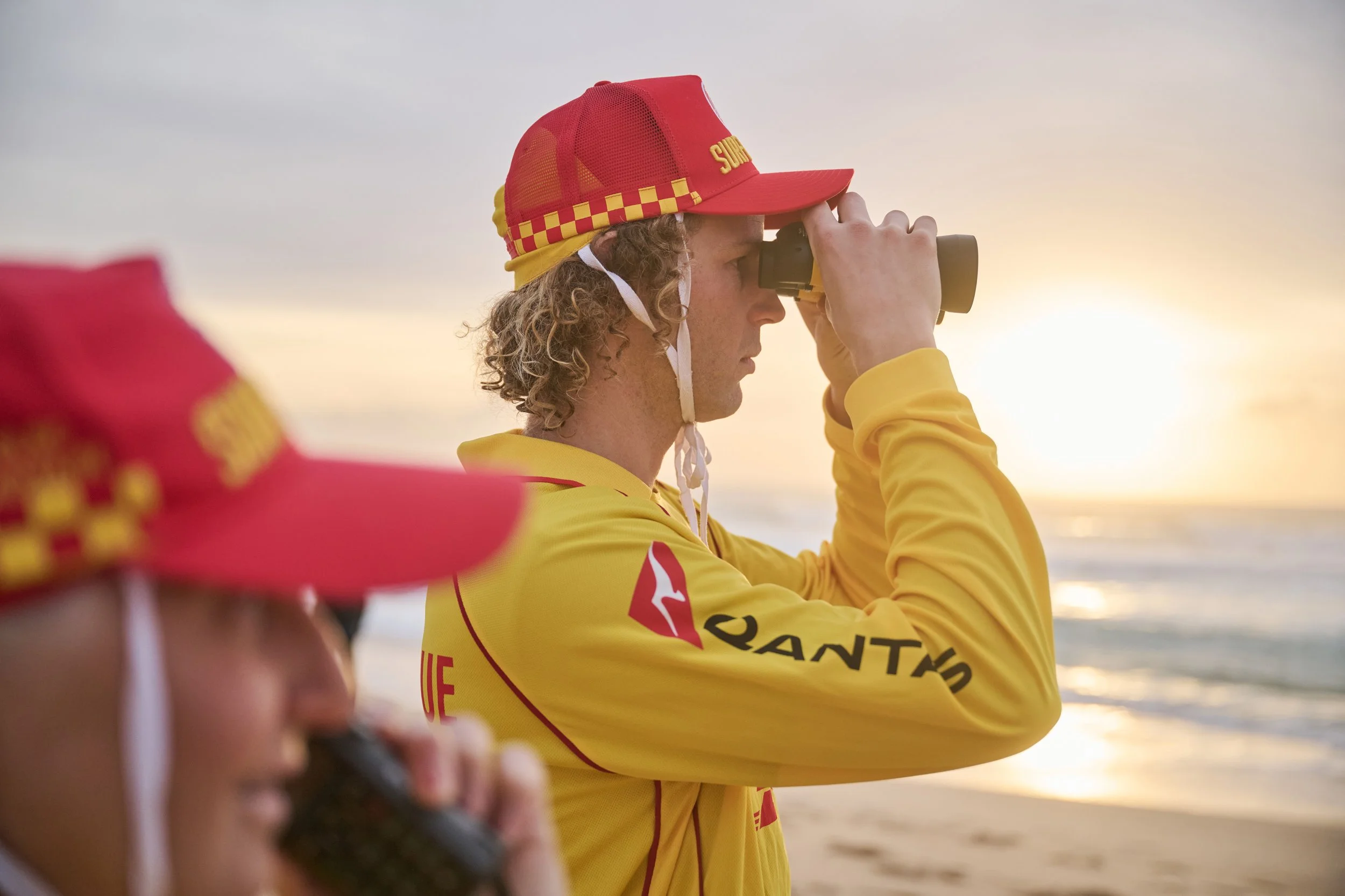 A young person in a yellow uniform and red cap with yellow checkered pattern, looking through binoculars at sunset, with another person blurred in the foreground.