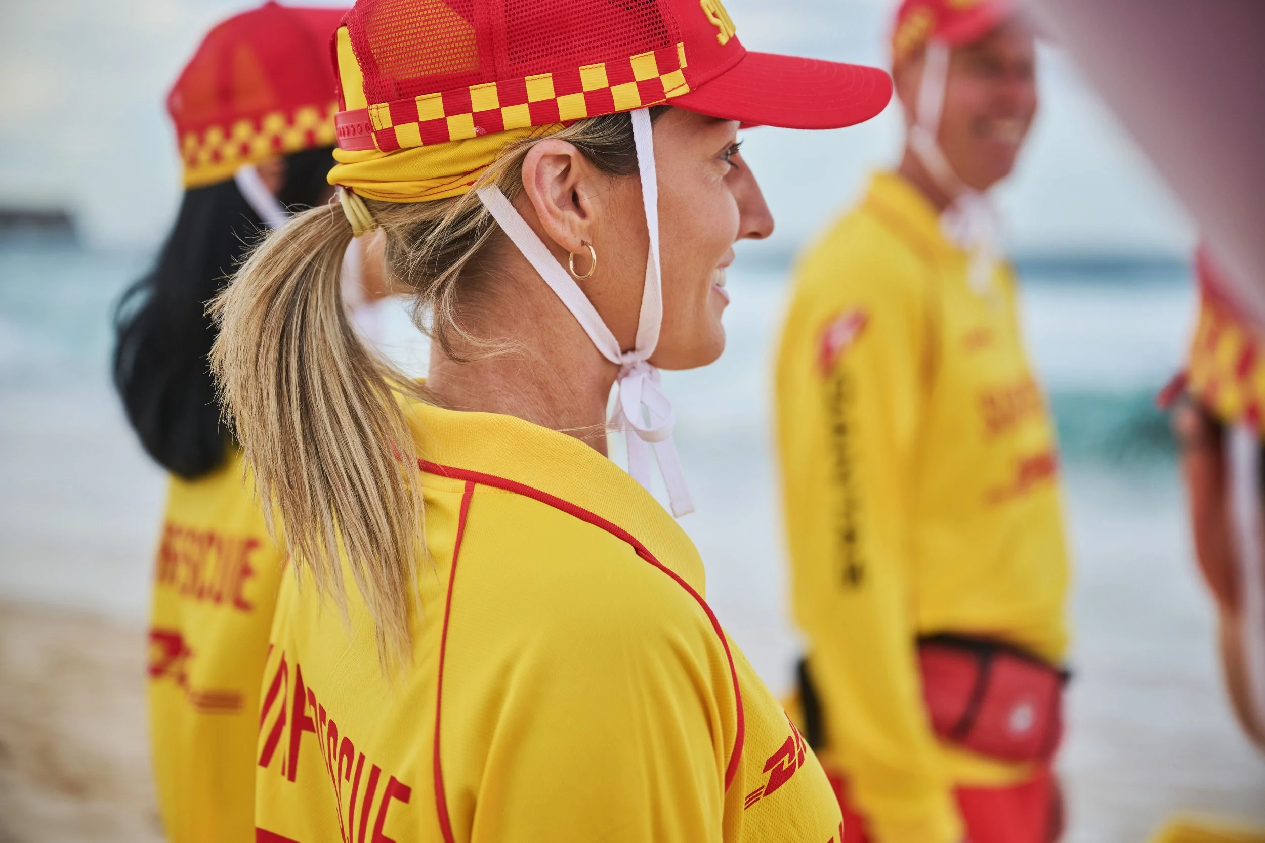 A woman wearing a red and yellow cap with a checkered pattern and a yellow jacket with red accents, standing on a beach with other women dressed similarly in the background.