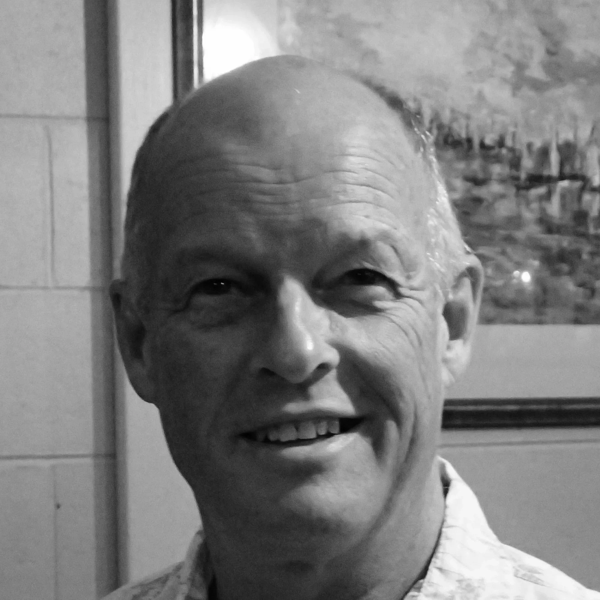 A black-and-white photo of a person's face, with wet hair, smiling, and looking at the camera, near a tiled wall and a doorframe.