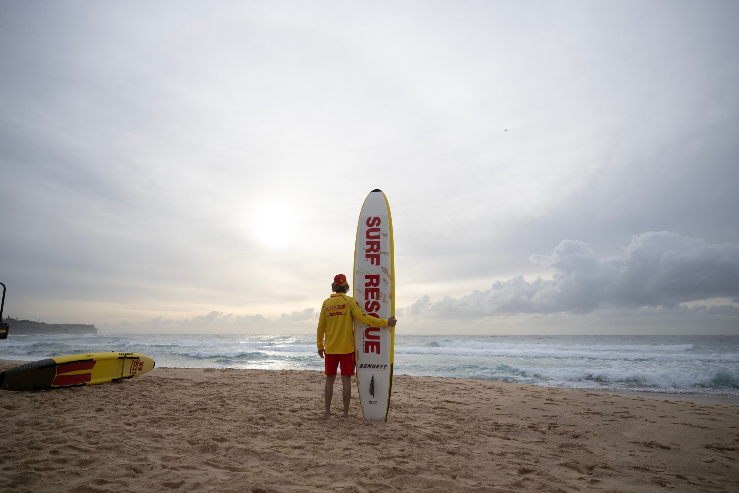 A surf rescue worker in a yellow jacket and red shorts holding a large life-saving board on a beach with ocean waves in the background and a cloudy sky.