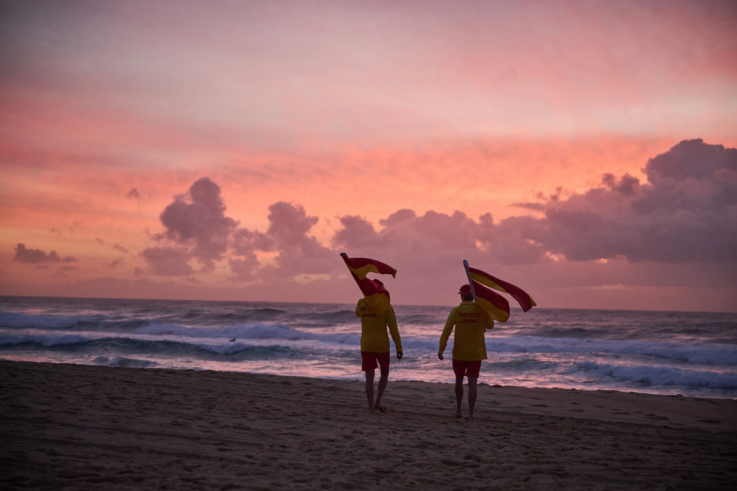 Beach scene at sunset with five rescue workers holding flags and walking along the shore, colorful sky with pink and purple clouds, ocean waves.