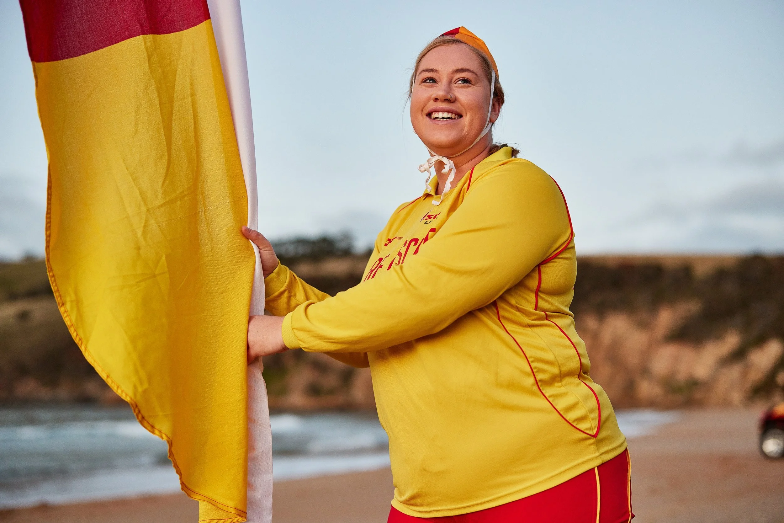 A smiling young woman in a yellow athletic jacket and red shorts stands outdoors, holding a yellow flag on a pole near a body of water during daytime.
