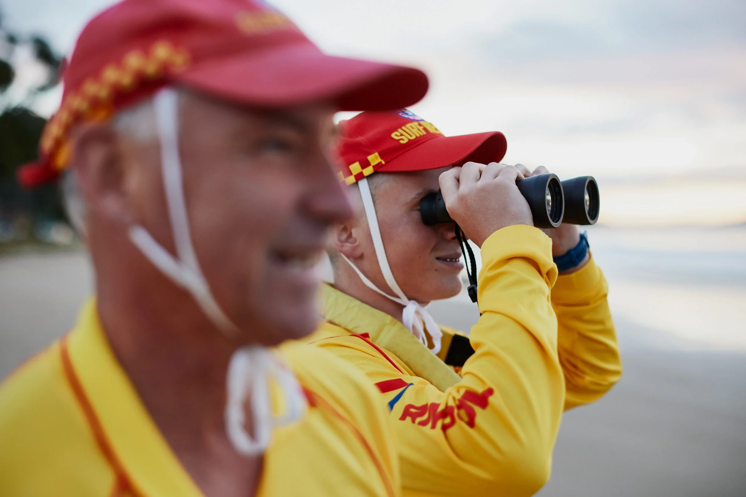 Two individuals in yellow shirts wearing red caps, one using binoculars, with a background of sky and possibly a beach or outdoor setting.
