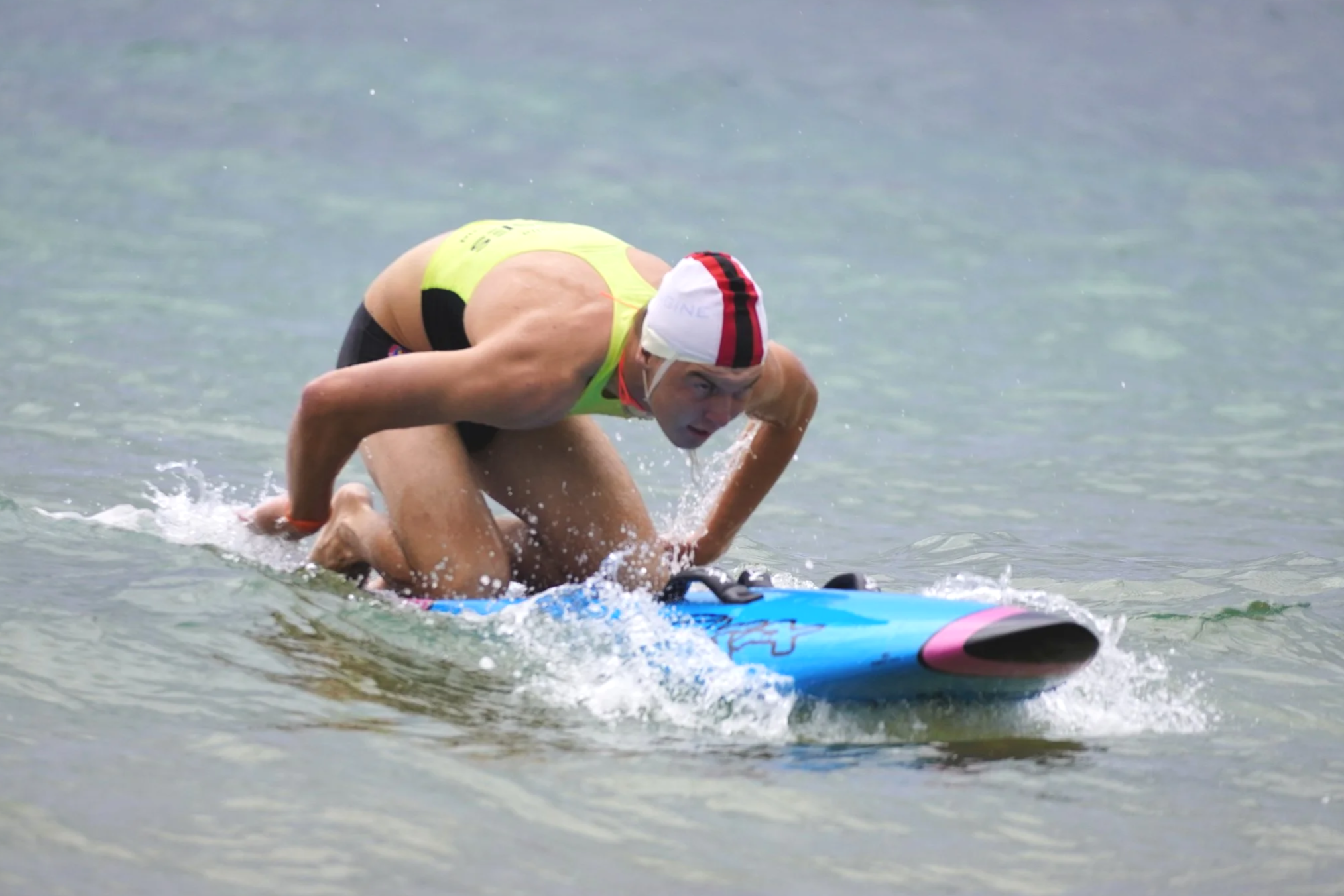 A man in a yellow swim suit and a red and white swim cap paddles on a surfboard in the water, leaning forward.