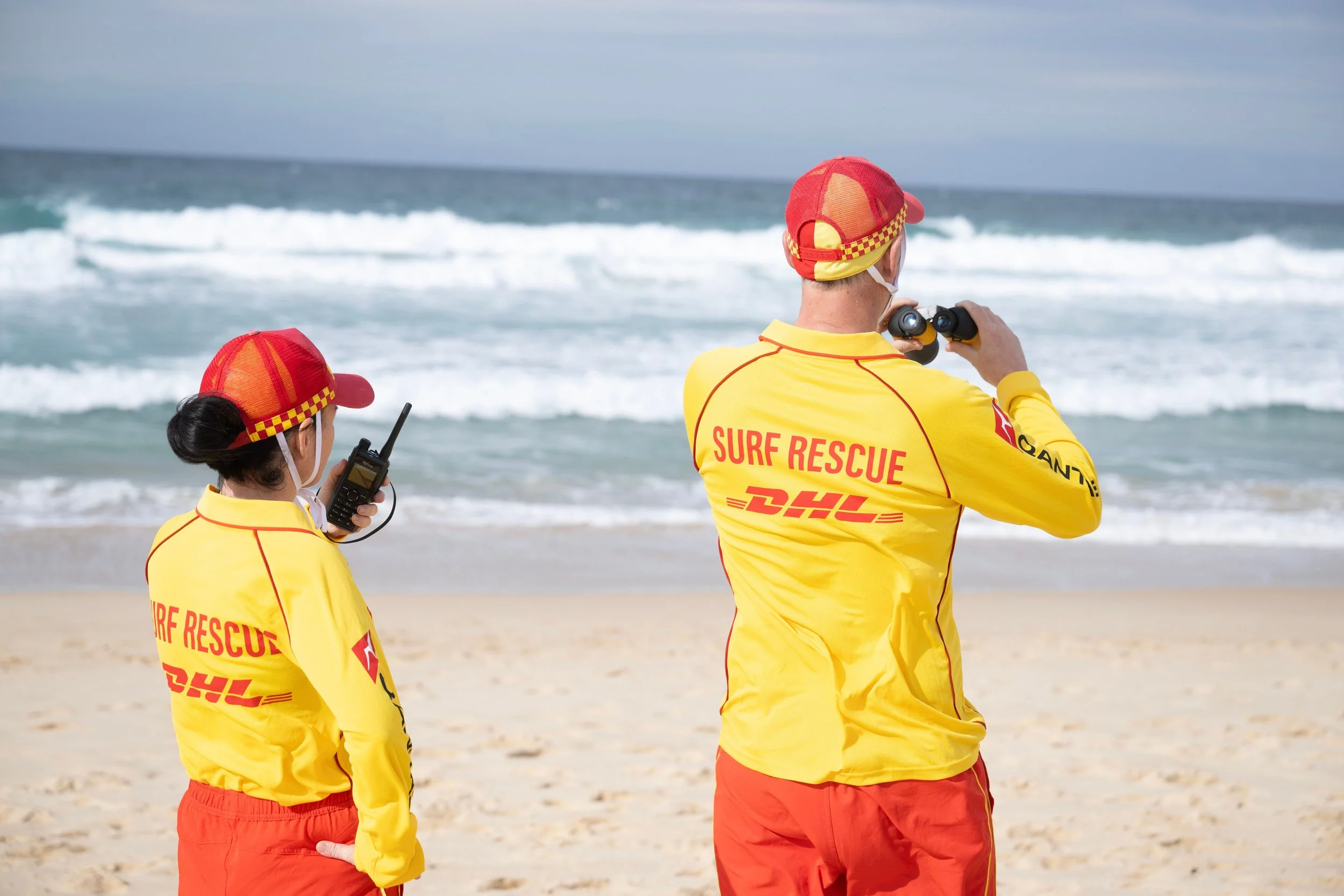 Two surf rescue team members in yellow shirts and red pants on the beach, looking out at the ocean with waves, one using binoculars and the other holding a radio.