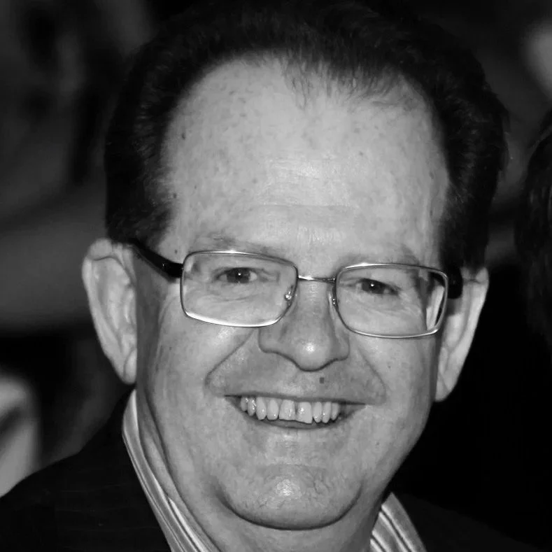 Black and white close-up photo of a smiling man with glasses, dark hair, and a collared shirt.