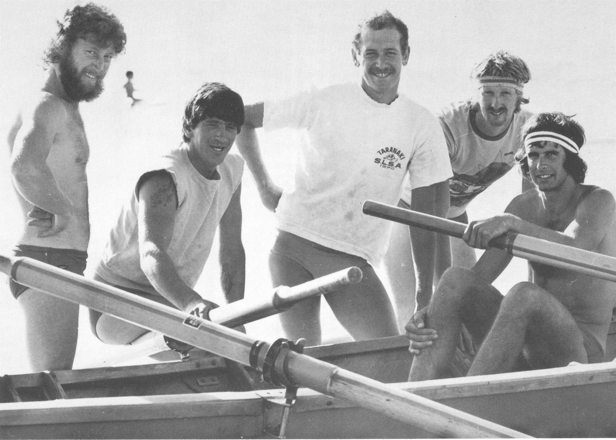 Five men are outdoors, smiling and gathered around a row boat, with one person in a white t-shirt and his hand on his hip.