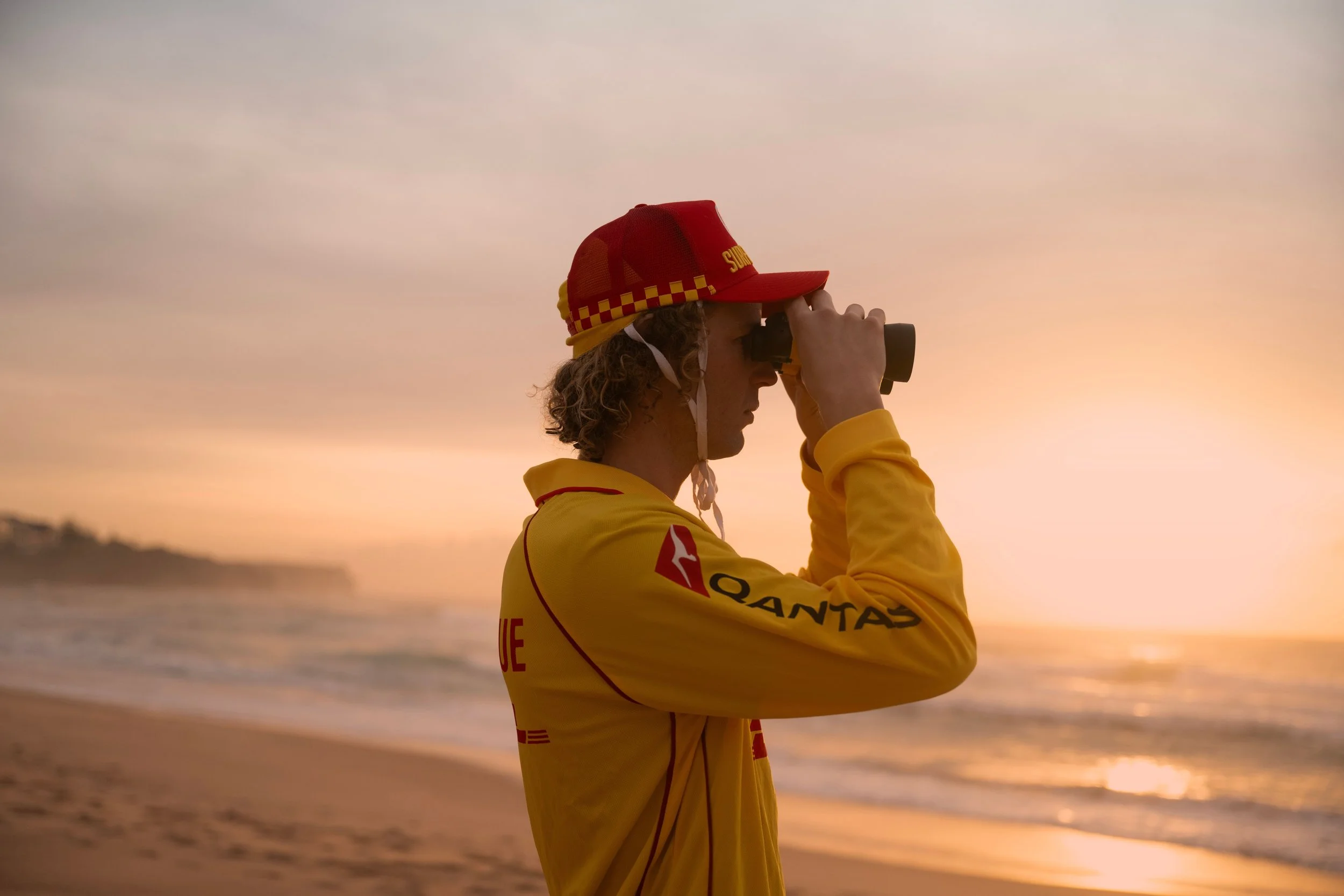 A person with curly hair wearing a red and yellow cap and a yellow jacket, looking through binoculars at the sunset by the water.