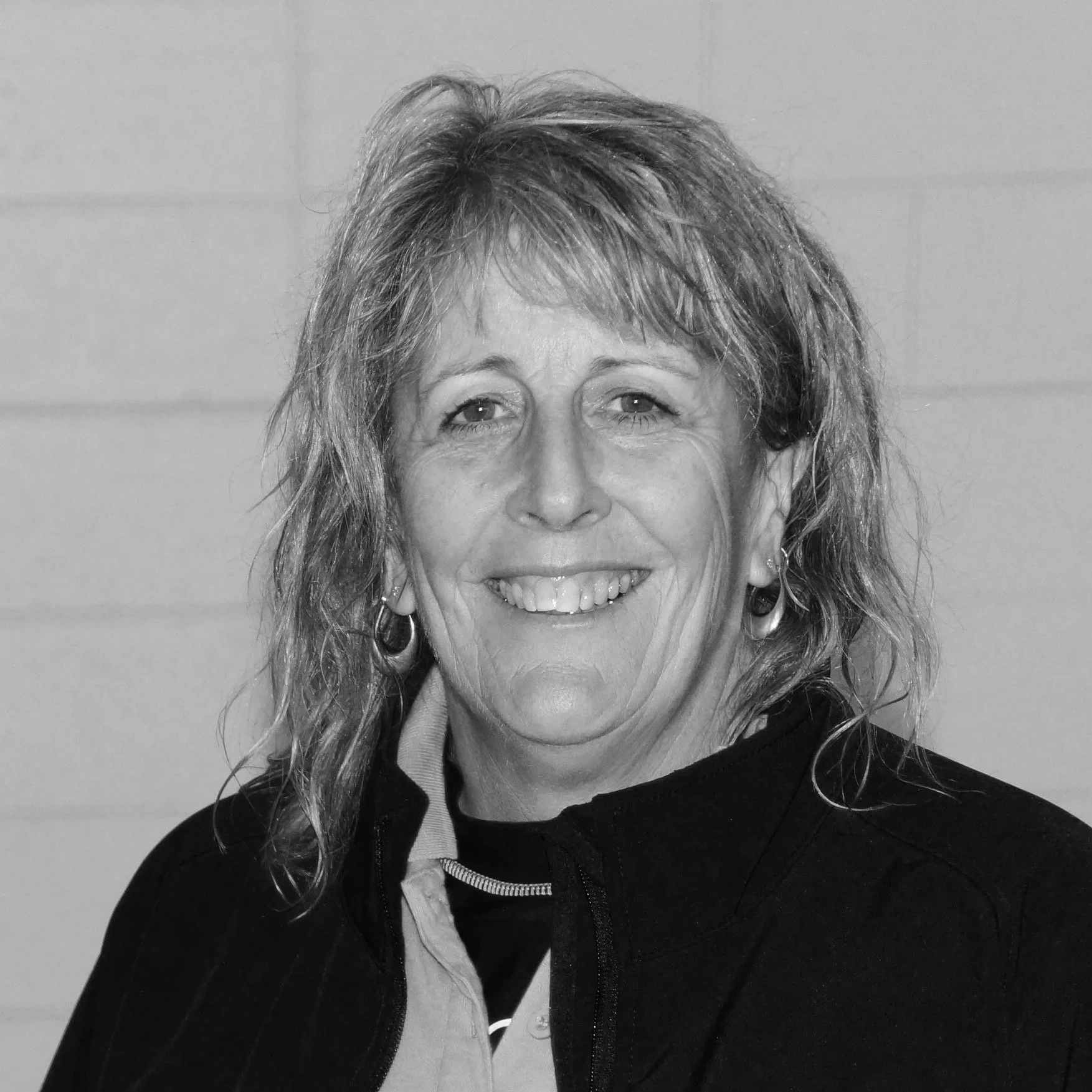 A woman with curly, shoulder-length hair smiling, wearing a black jacket over a light-colored shirt, and earrings, in front of a plain background.