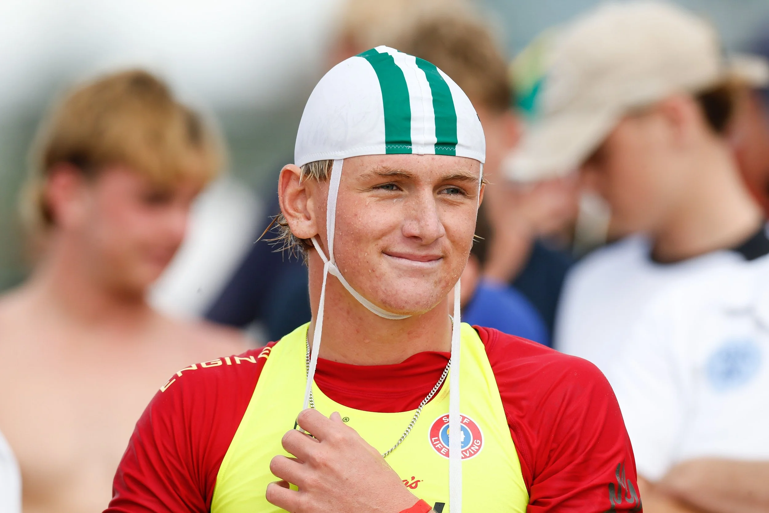 Close-up of a young male athlete with wet skin, wearing a white swimming cap with green stripes and a red and yellow sports uniform, standing among others at a sports event.