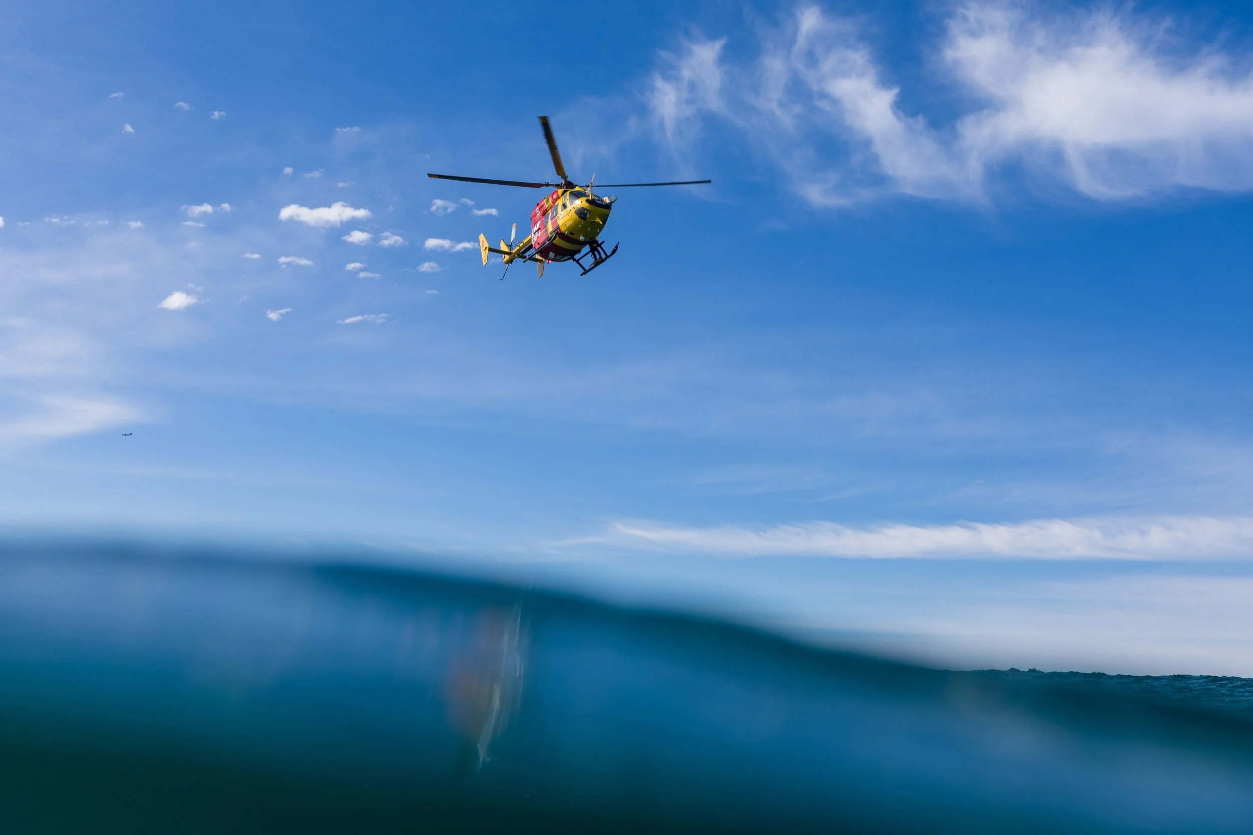 A yellow and red rescue helicopter flying over a body of water under a blue sky with scattered clouds.