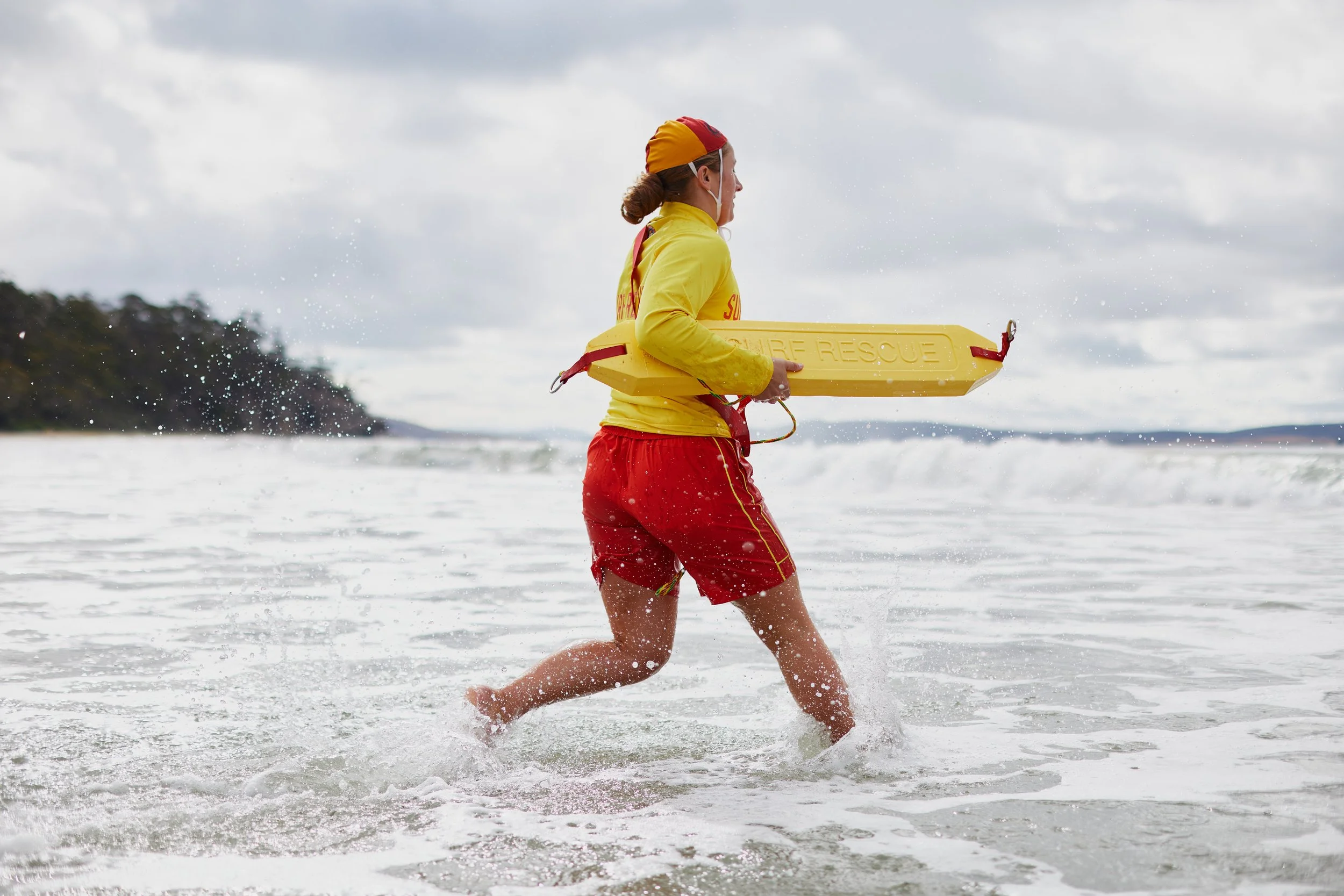 A lifeguard in red shorts and a yellow shirt carrying a yellow rescue float while running into the ocean during cloudy weather.