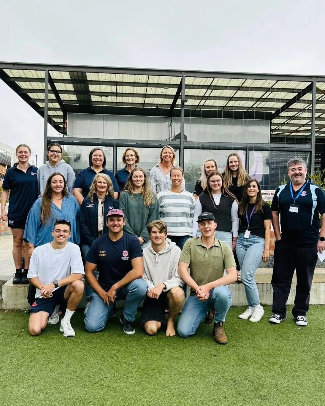 Group of young adults, teenagers, and adults smiling and posing for a photo outdoors in front of a modern building with a glass and metal facade. The group is arranged in two rows, with some kneeling in front and others standing behind.