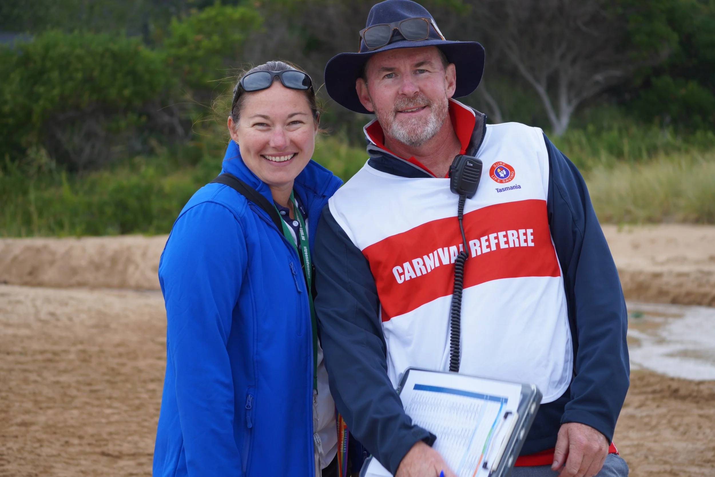A woman and a man at the beach, both smiling and wearing casual outdoor clothing. The man is wearing a wide-brim hat, sunglasses, and a shirt with a badge that says 'CARNIVAL REFEREE,' and is holding a clipboard. The woman is wearing sunglasses on her head and a blue jacket.