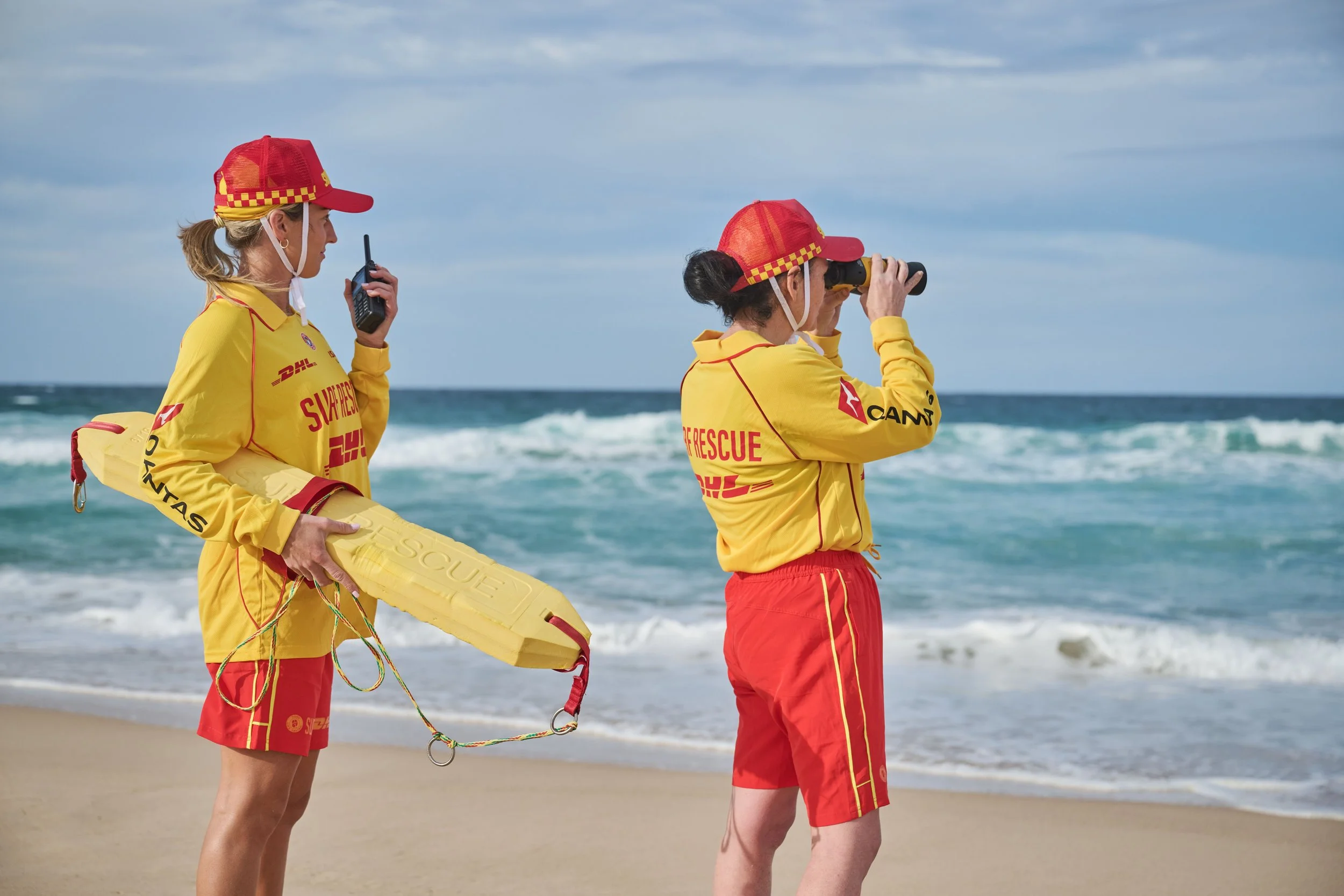 Two female lifeguards on a beach, wearing yellow and red rescue uniforms and hats, one holding a radio and the other looking through binoculars, with ocean waves in the background.