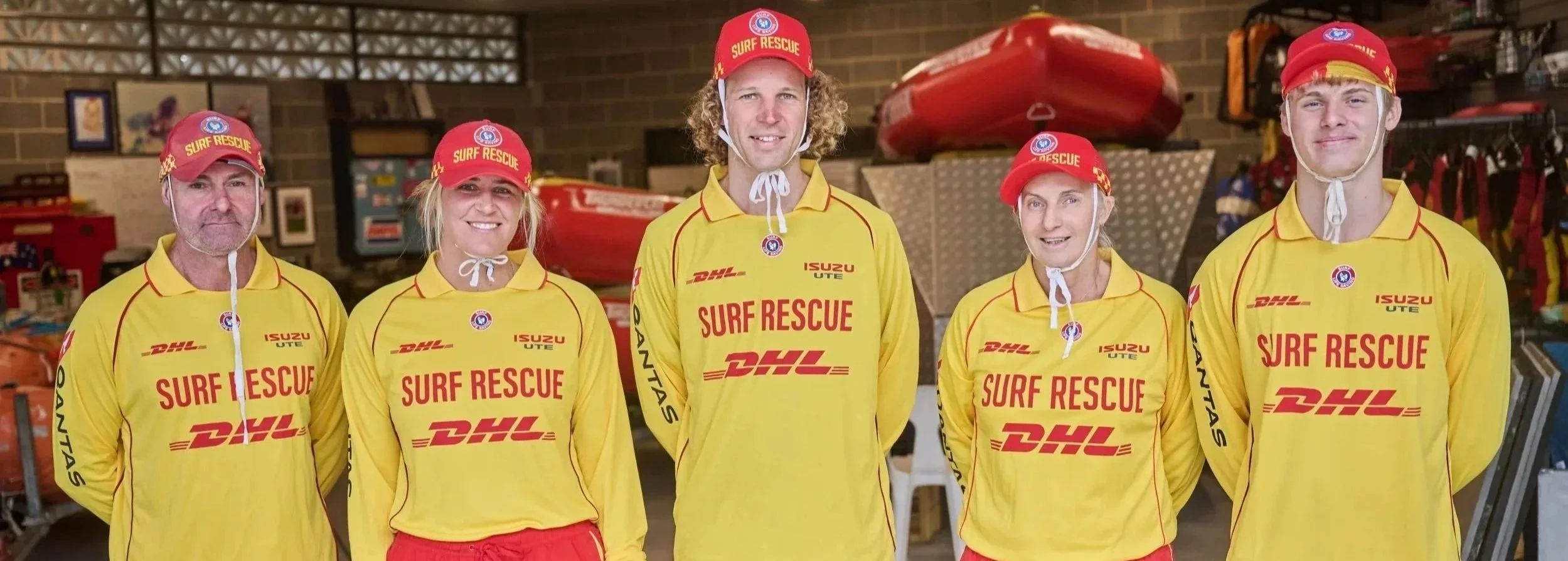 Six surf rescue team members standing indoors, wearing yellow uniforms with logos, red caps labeled 'SURF RESCUE', and standing in front of rescue equipment and boat.
