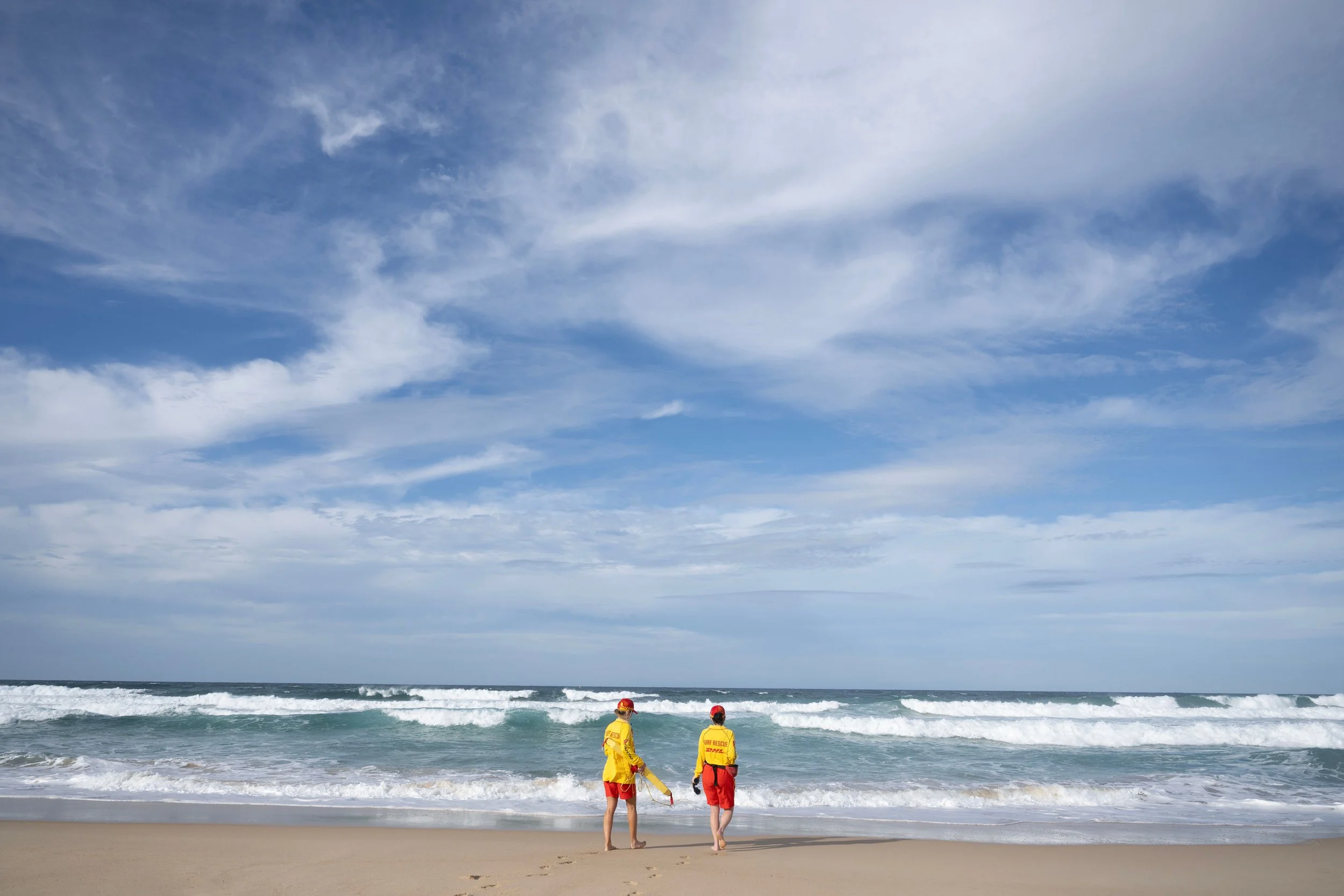 Two lifeguards in yellow uniforms with red accents standing on the sandy beach facing the ocean, which has gentle waves and a partly cloudy sky above.