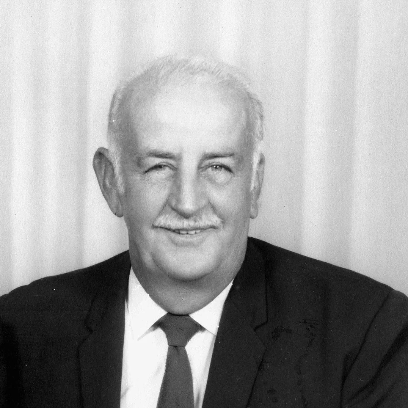 Black and white portrait of an older man with light-colored hair and a mustache, wearing a suit and tie, smiling at the camera.