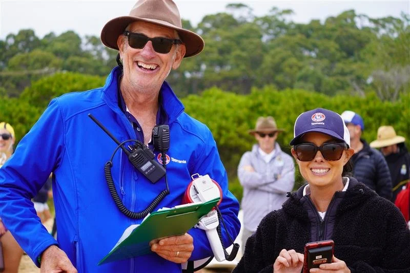 A group of people outdoors, smiling. The man in the foreground is wearing a wide-brimmed hat, sunglasses, a blue jacket, and is holding a clipboard and radio. The woman next to him is wearing sunglasses, a cap, and is holding a phone. Other people are standing in the background, also wearing hats and sunglasses.