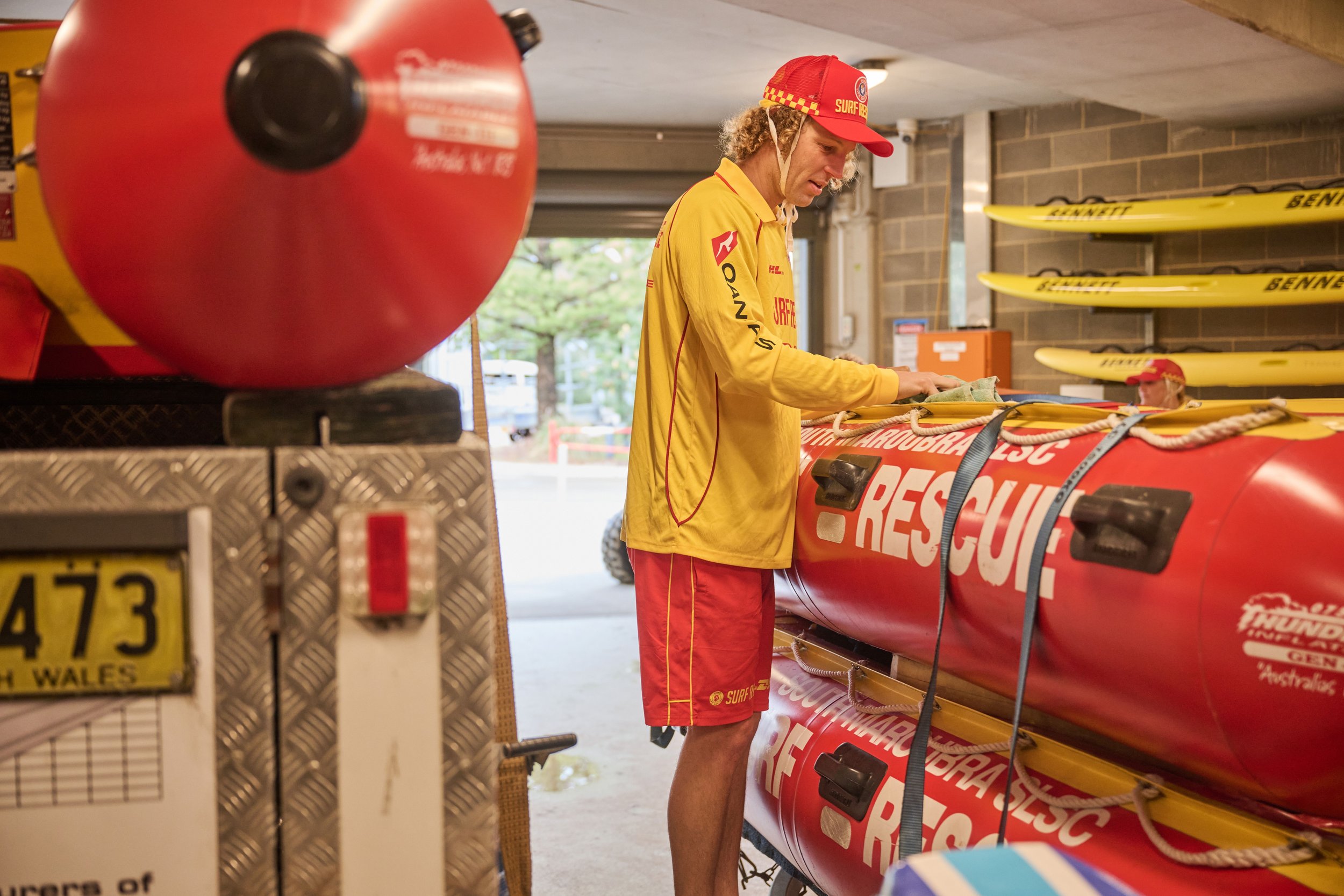 A member of a rescue team wearing a yellow shirt and red cap is working with equipment in a storage area, with other rescue personnel in the background.