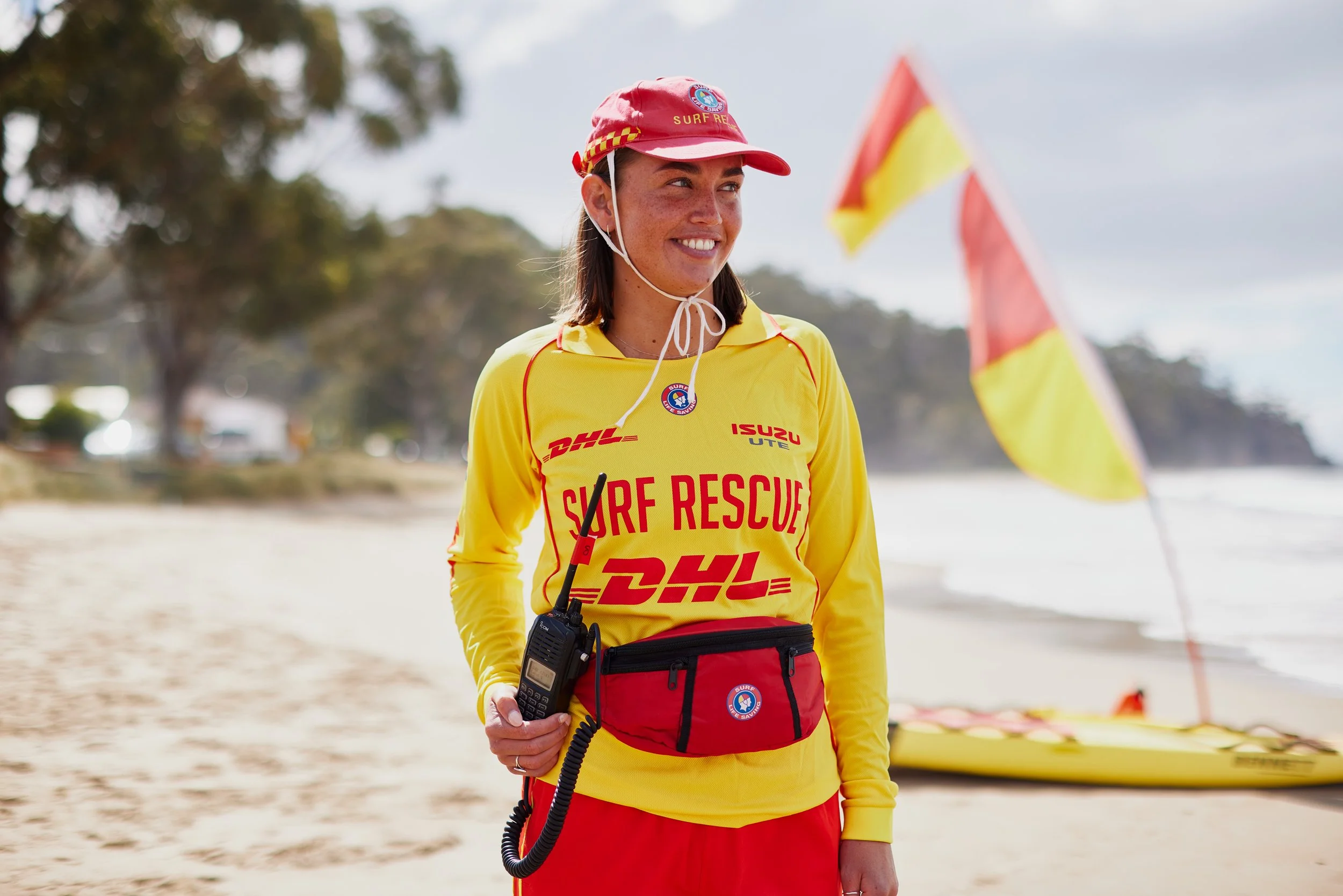 A woman wearing a yellow surf rescue uniform, a red cap, and a radio, standing on the beach near a yellow rescue surfboard and flags.