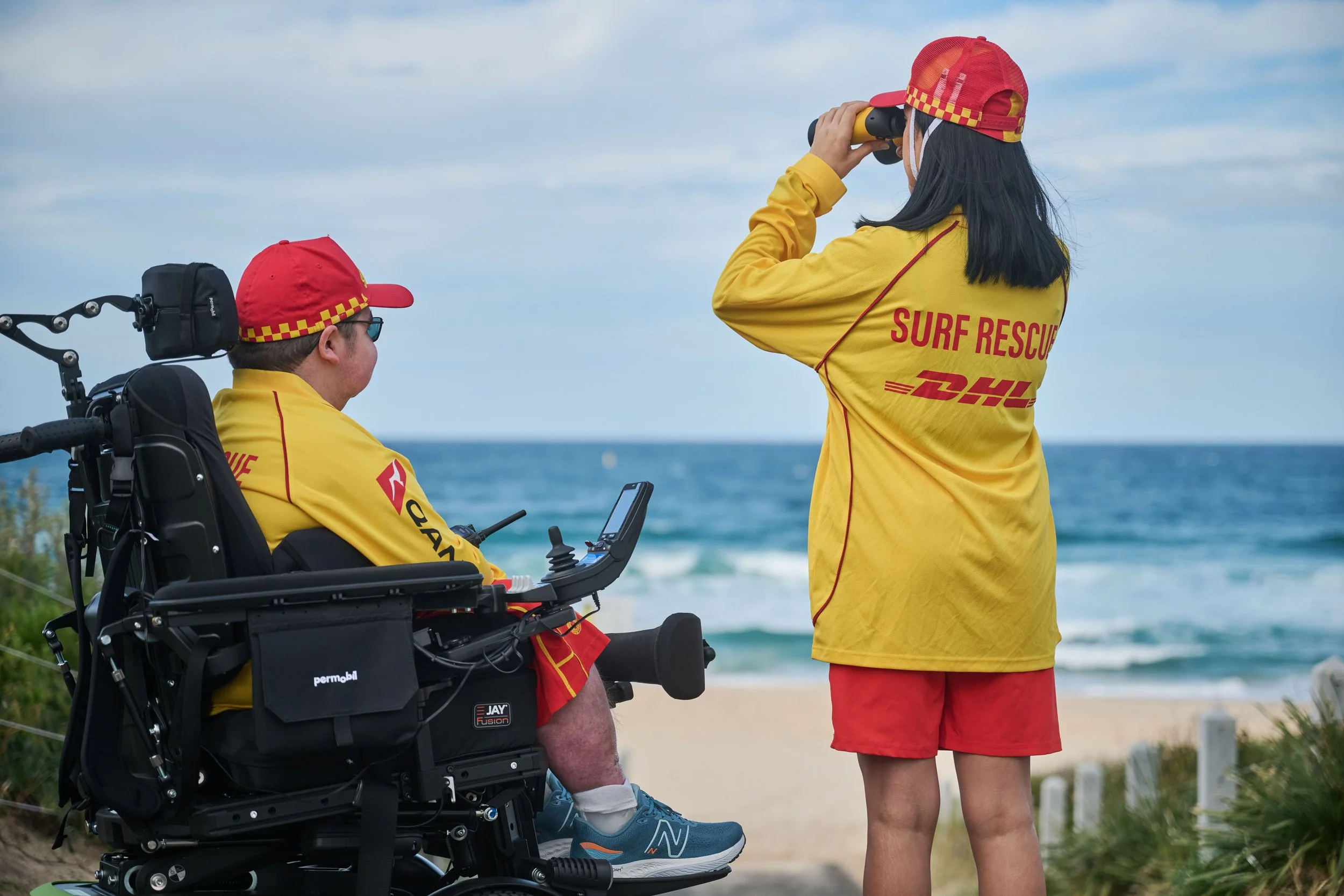 A person in a wheelchair and a person standing on a beach. The person in the wheelchair is wearing a yellow surf rescue shirt and a red cap. The standing person is dressed in a yellow surf rescue shirt with a DHL logo, red shorts, and a red cap with a checkered yellow pattern. The person standing is holding binoculars and looking at the ocean, with the sky and ocean visible in the background.
