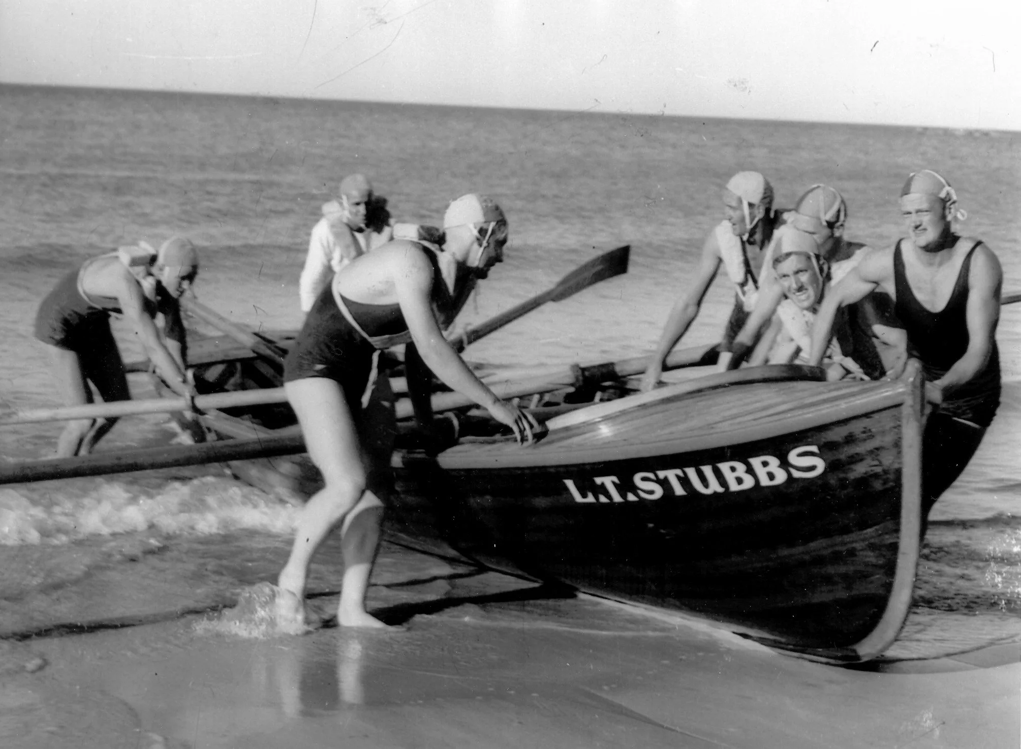 Black and white photo of a group of people on the beach preparing a small boat for launching, some wearing swim caps and wetsuits, with the sea in the background.
