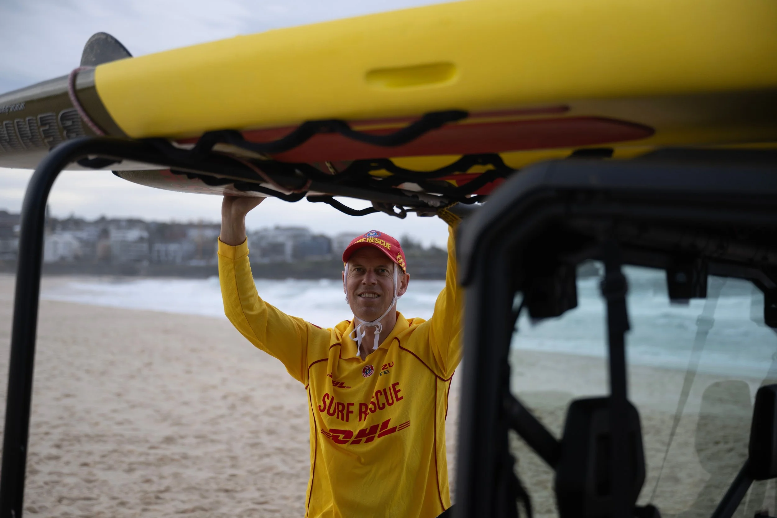 A person wearing a yellow surf rescue uniform and a red cap is standing on the beach, holding a yellow rescue board above their head.