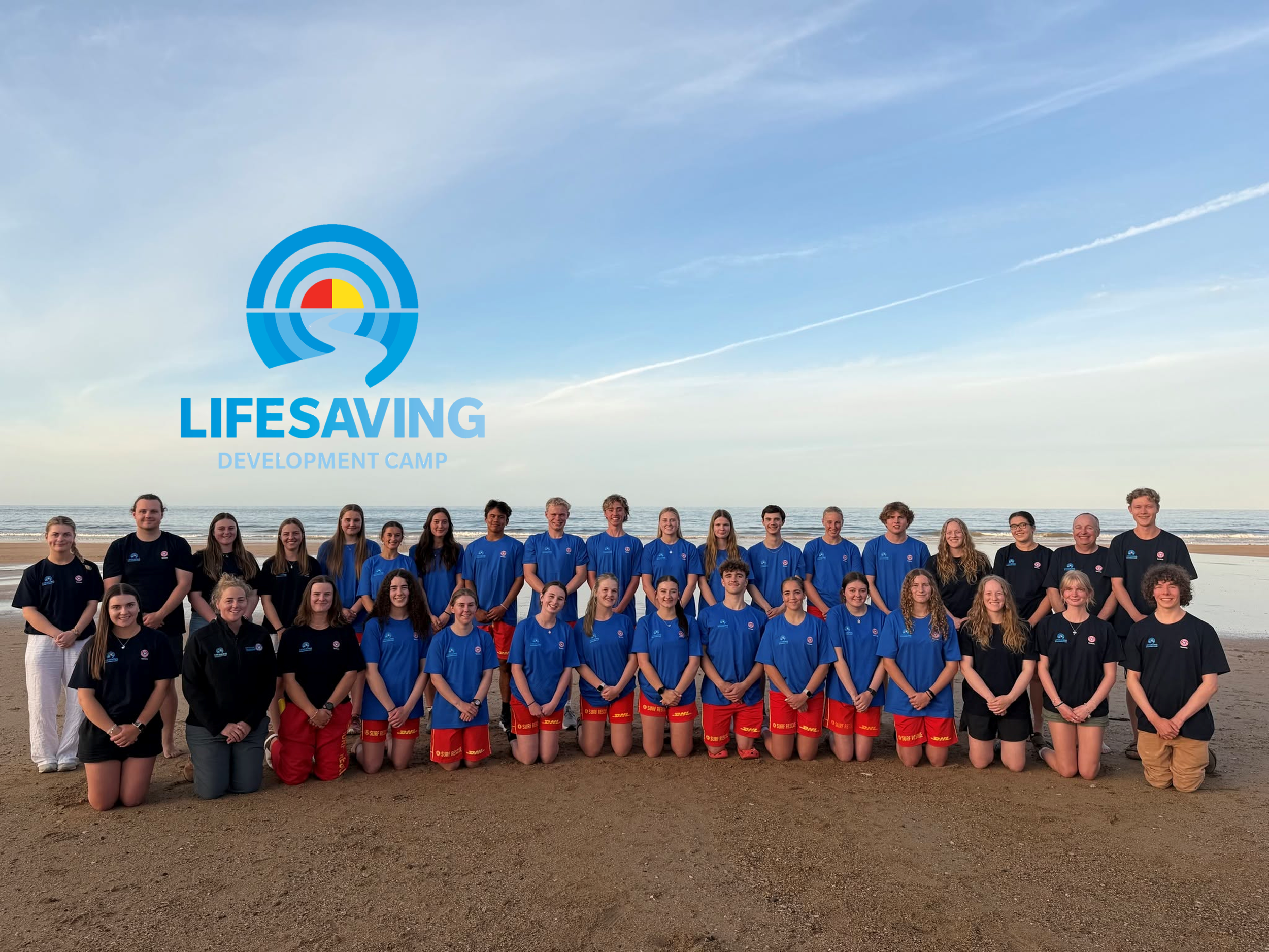 A large group of people, primarily young adults, are standing on a beach during the day, with the ocean and sky in the background. They are dressed in matching shirts and shorts, likely participating in a lifeguarding or beach safety development camp.