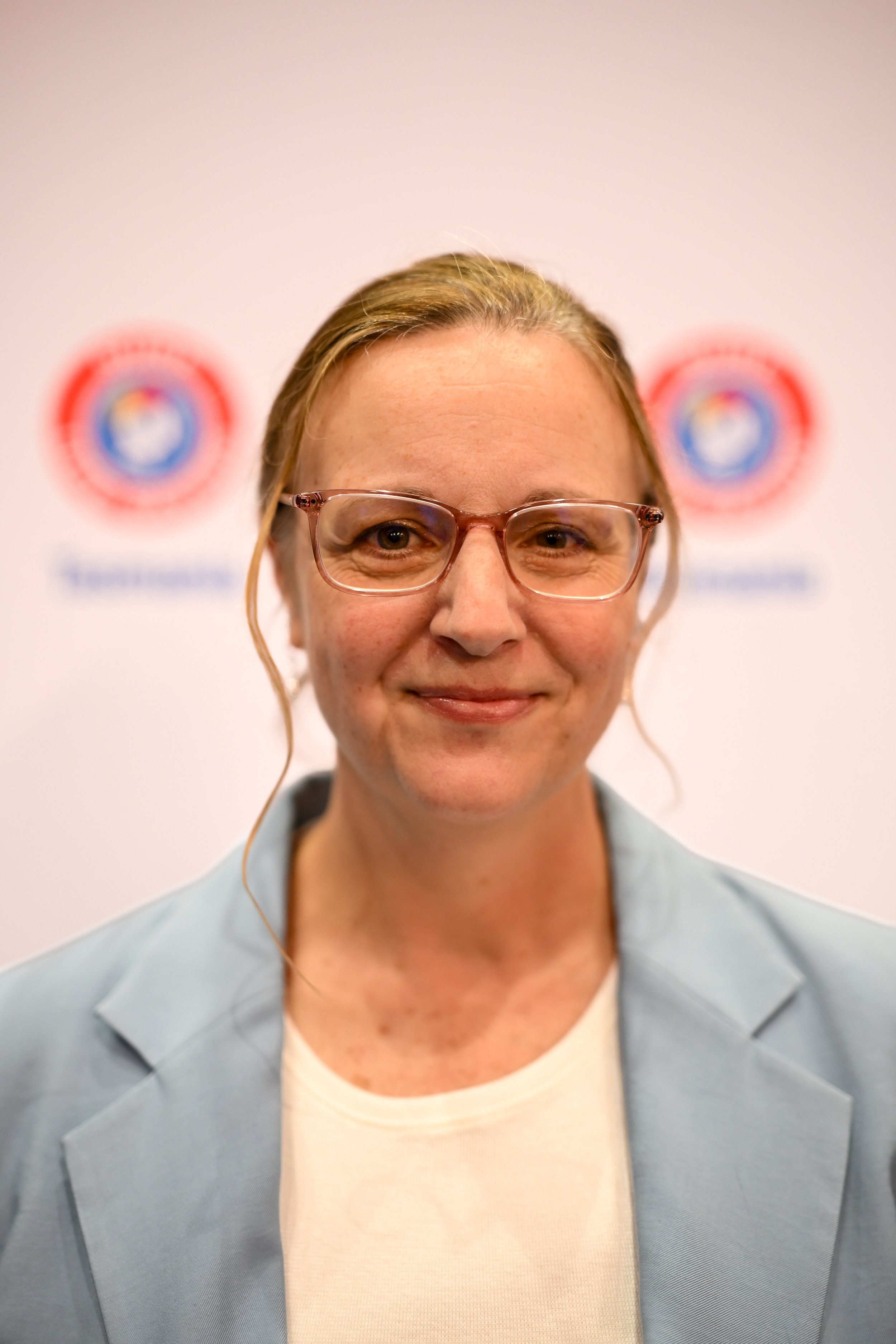 Close-up of a woman with glasses smiling, wearing a light gray blazer and a white shirt, standing in front of a backdrop with logos that are blurred.