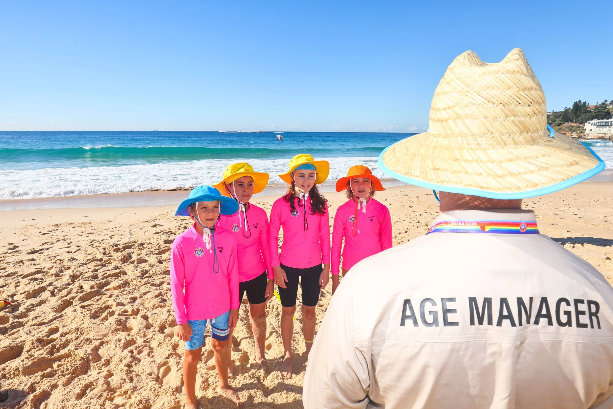 Four children dressed in pink swimsuits and colorful sun hats standing on a sandy beach near the ocean, with a person in a white shirt labeled 'Age Manager' in the foreground and a large straw hat in the lower right corner.