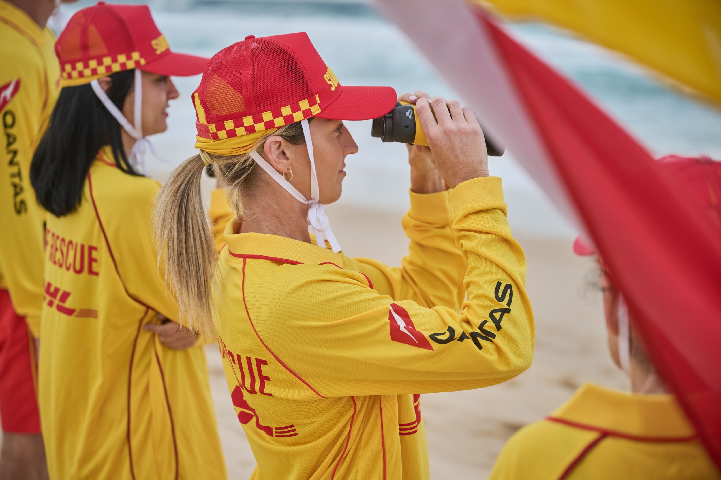Women wearing yellow rescue uniforms and red hats with checkered pattern, one using binoculars at the beach.