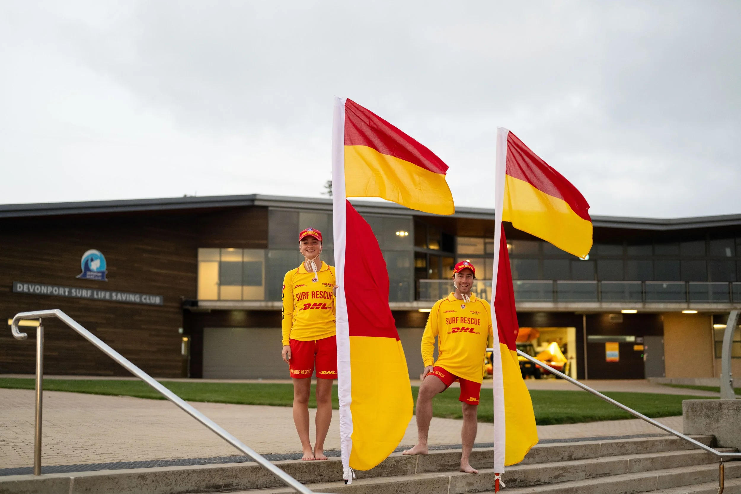 Three surf rescue team members standing outside of a surf club building, holding red and yellow flags. They are wearing yellow uniforms with red accents and safety hats. The building has a sign that reads 'Devonport Surf Life Saving Club'. It is cloudy and they are on steps next to a railing.