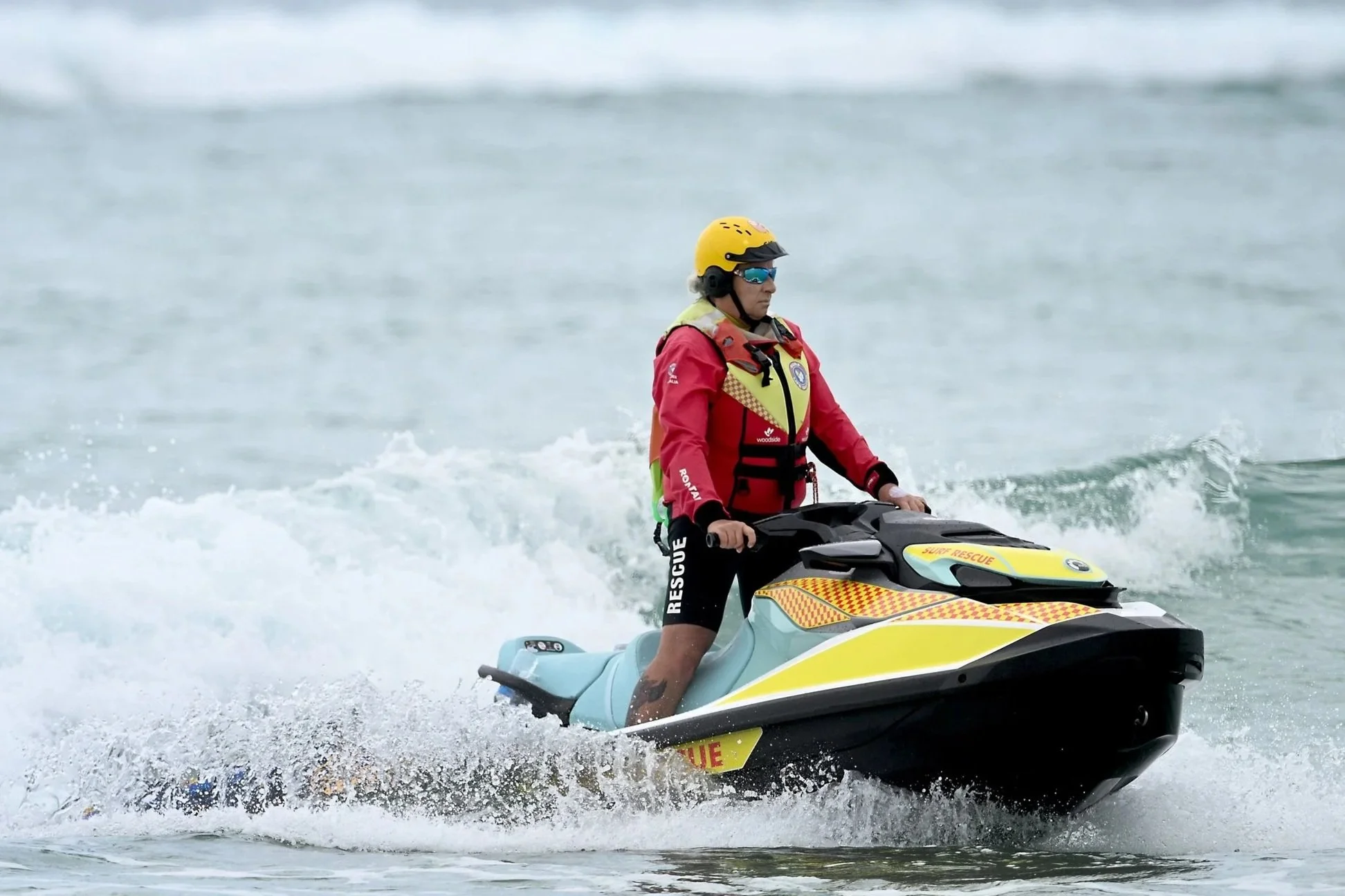 A lifeguard riding a jet ski on the water, wearing a yellow helmet, sunglasses, a red rescue jacket, and black shorts with 'RESCUE' printed on them.