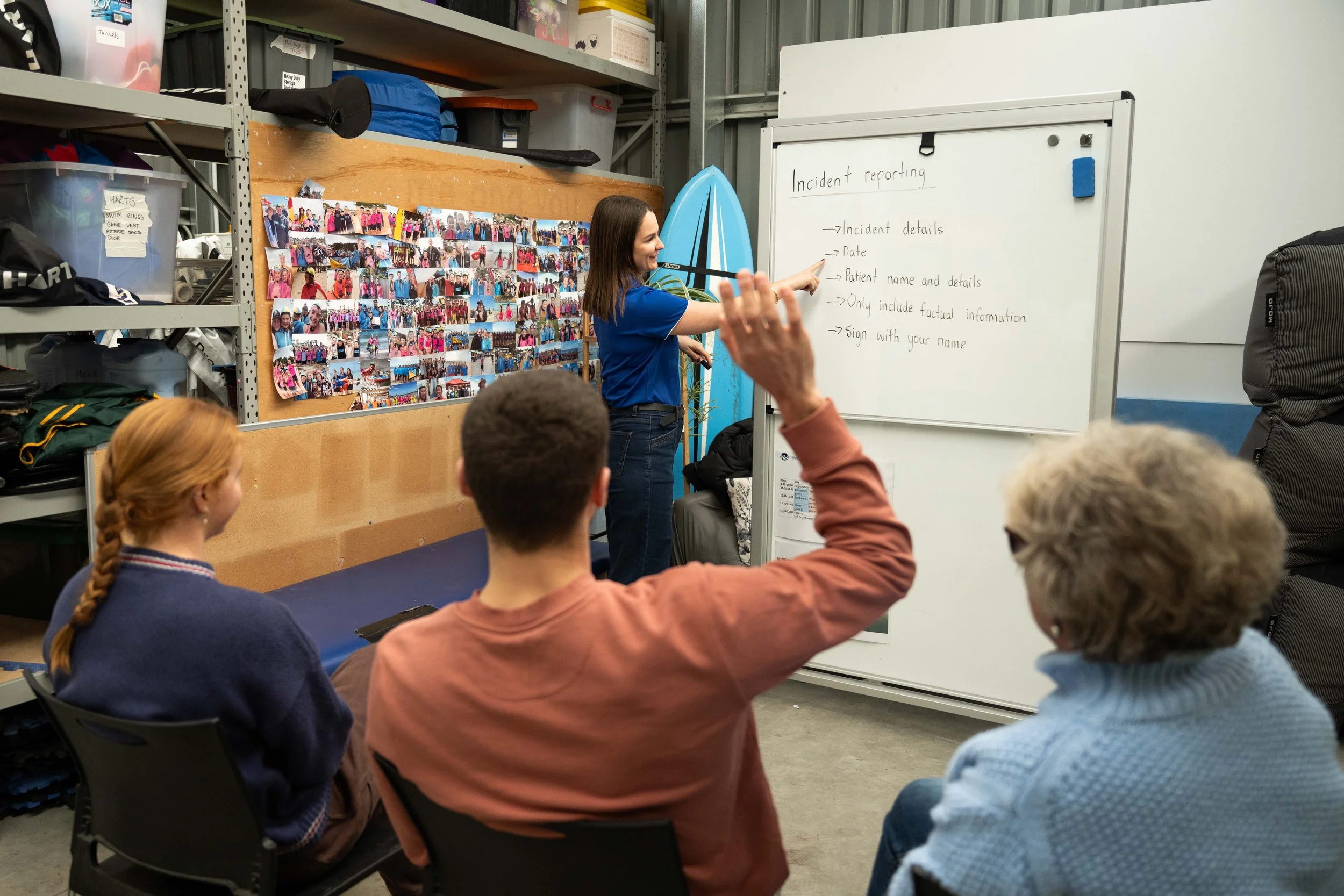 A woman in blue shirt giving a presentation to a group of seated people in a storage room with a whiteboard that has incident reporting tips, and a wall covered with photographs of group activities.