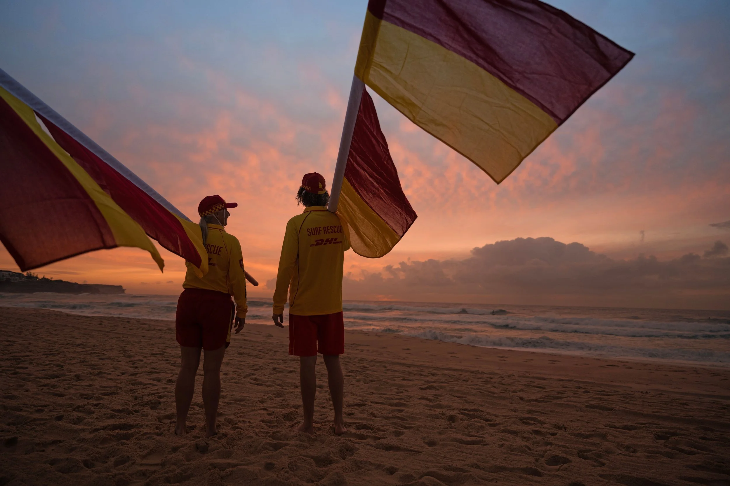 Two surf rescue workers in yellow and red uniforms and caps on a beach at sunset, holding large flags with the ocean in the background.