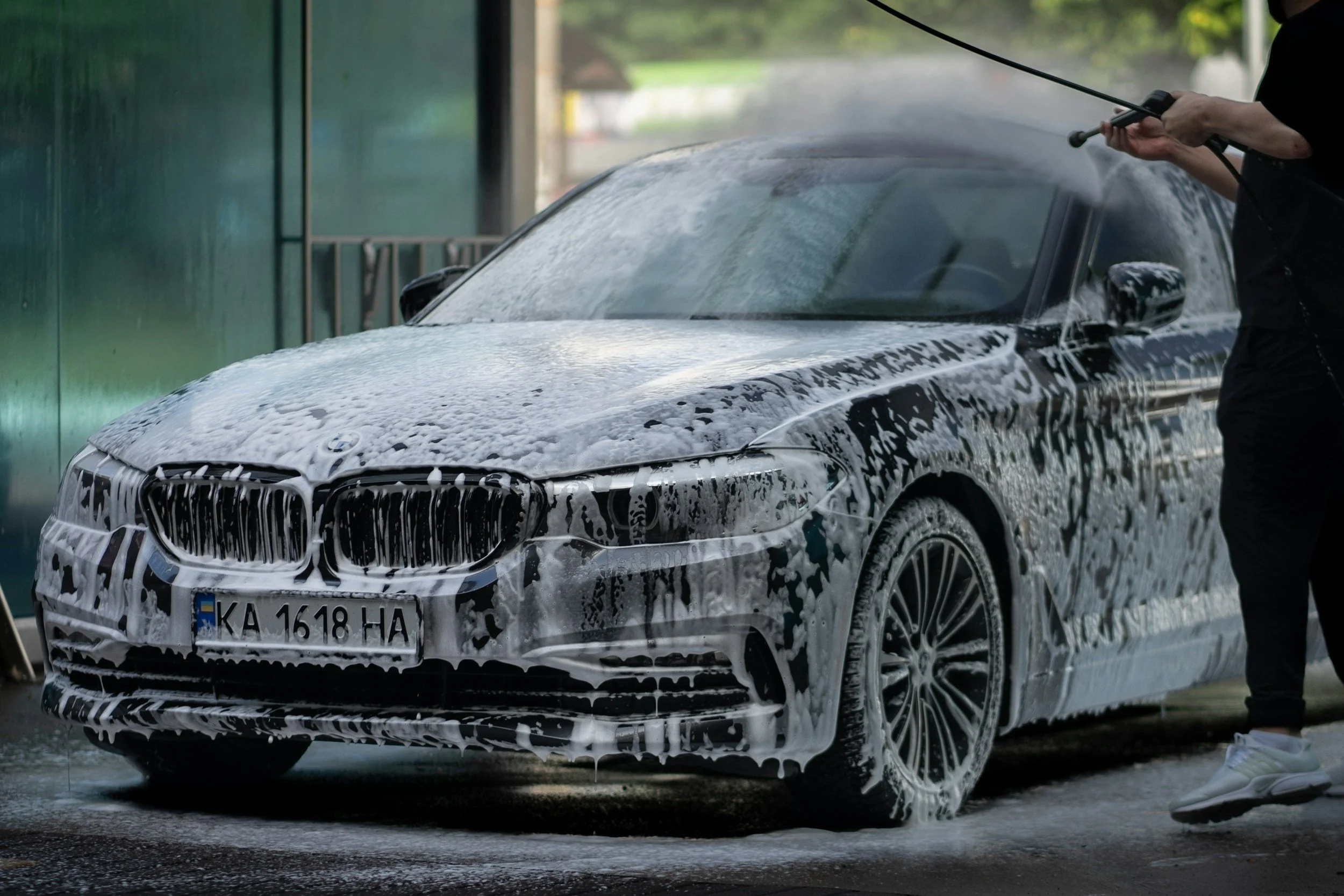 A person cleaning a black BMW car with soap and water, with foam covering the front and side, during daytime.