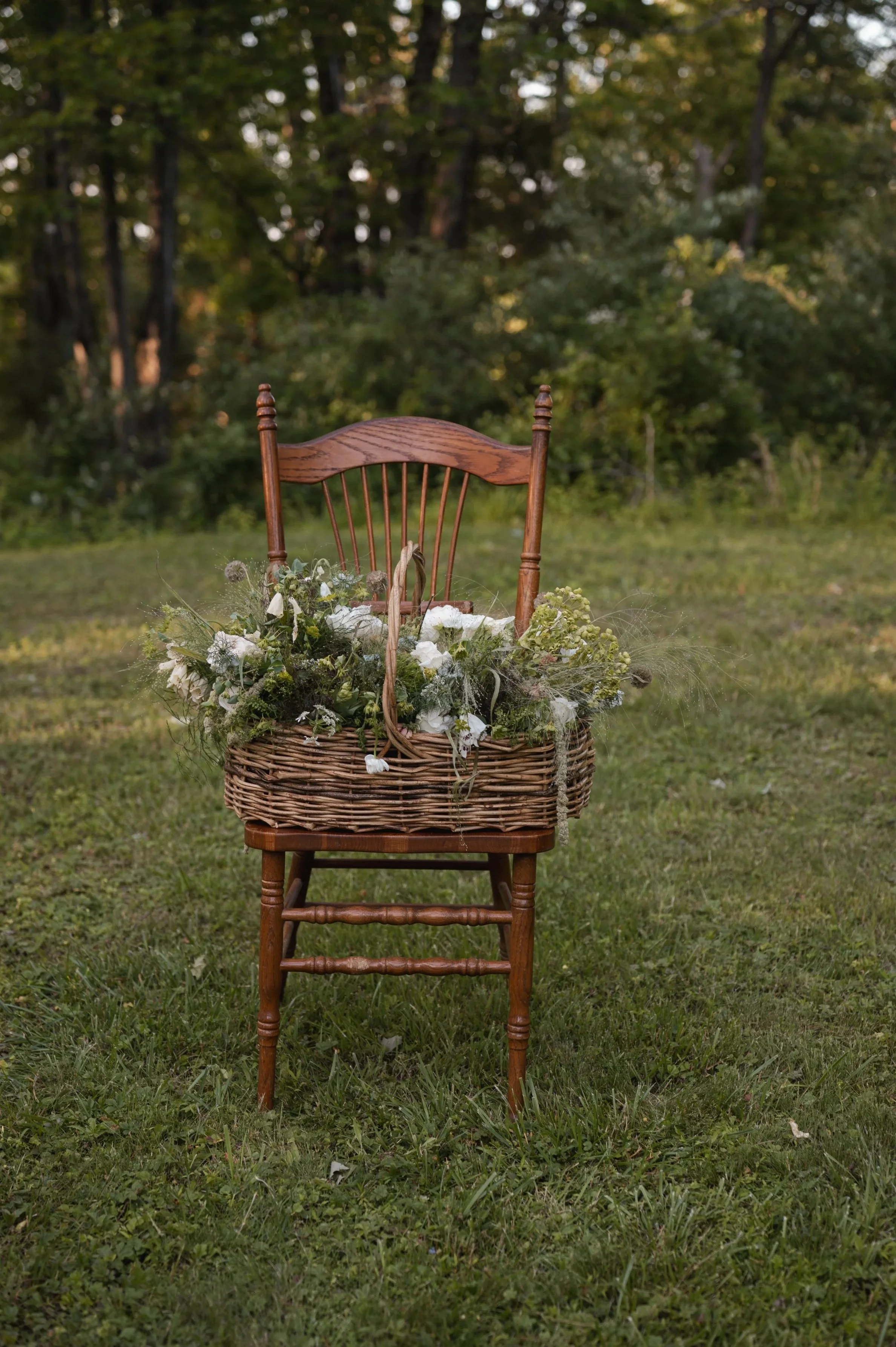 A wooden chair with a woven basket filled with white and green flowers placed on its seat, outdoors on grass with a background of trees and foliage.