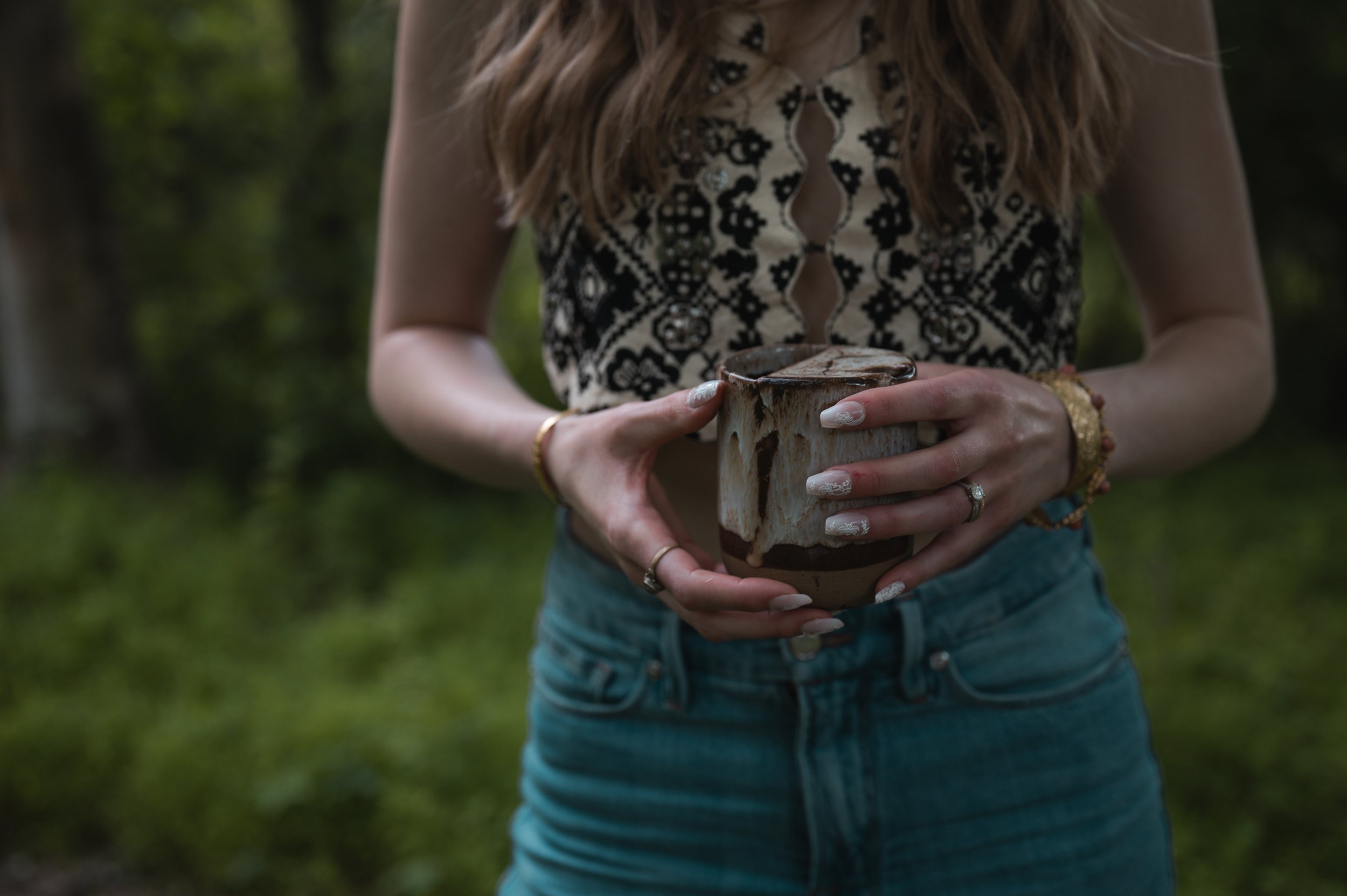 A woman in a sleeveless top with a black and white pattern is holding a ceramic cup with a liquid, likely coffee or hot chocolate, outdoors in a forested area.