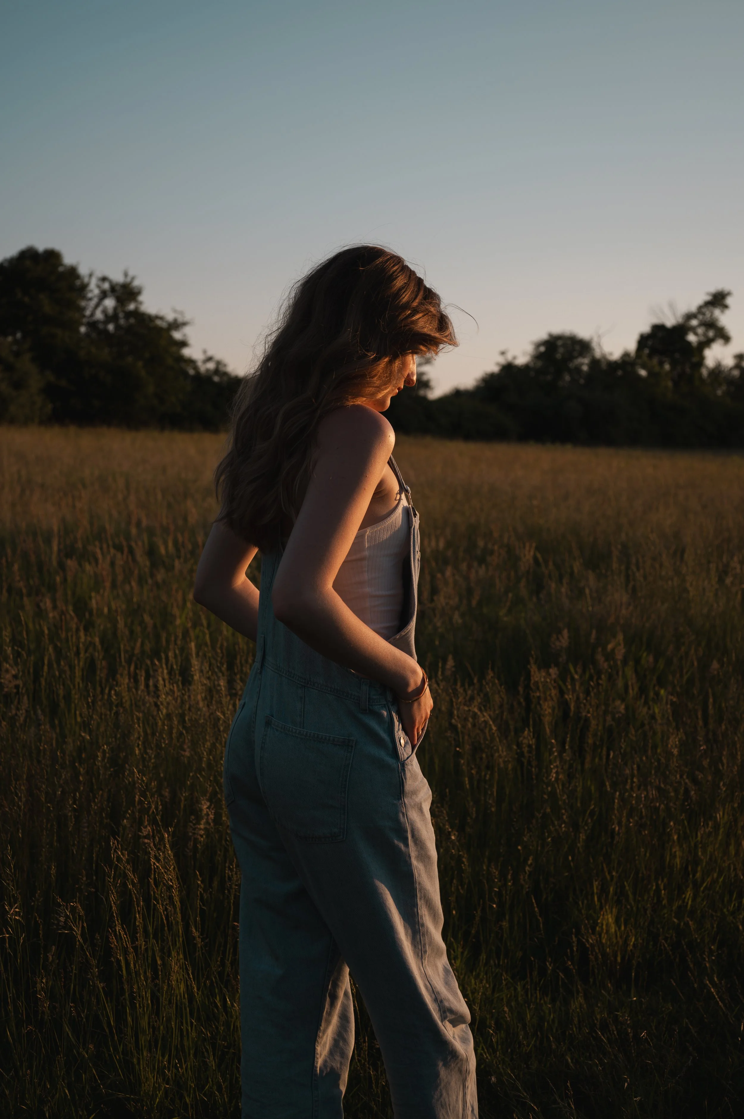 A woman with long brown hair in a flowing style is standing in a field of tall grass at sunset, with trees in the background and a clear sky overhead.