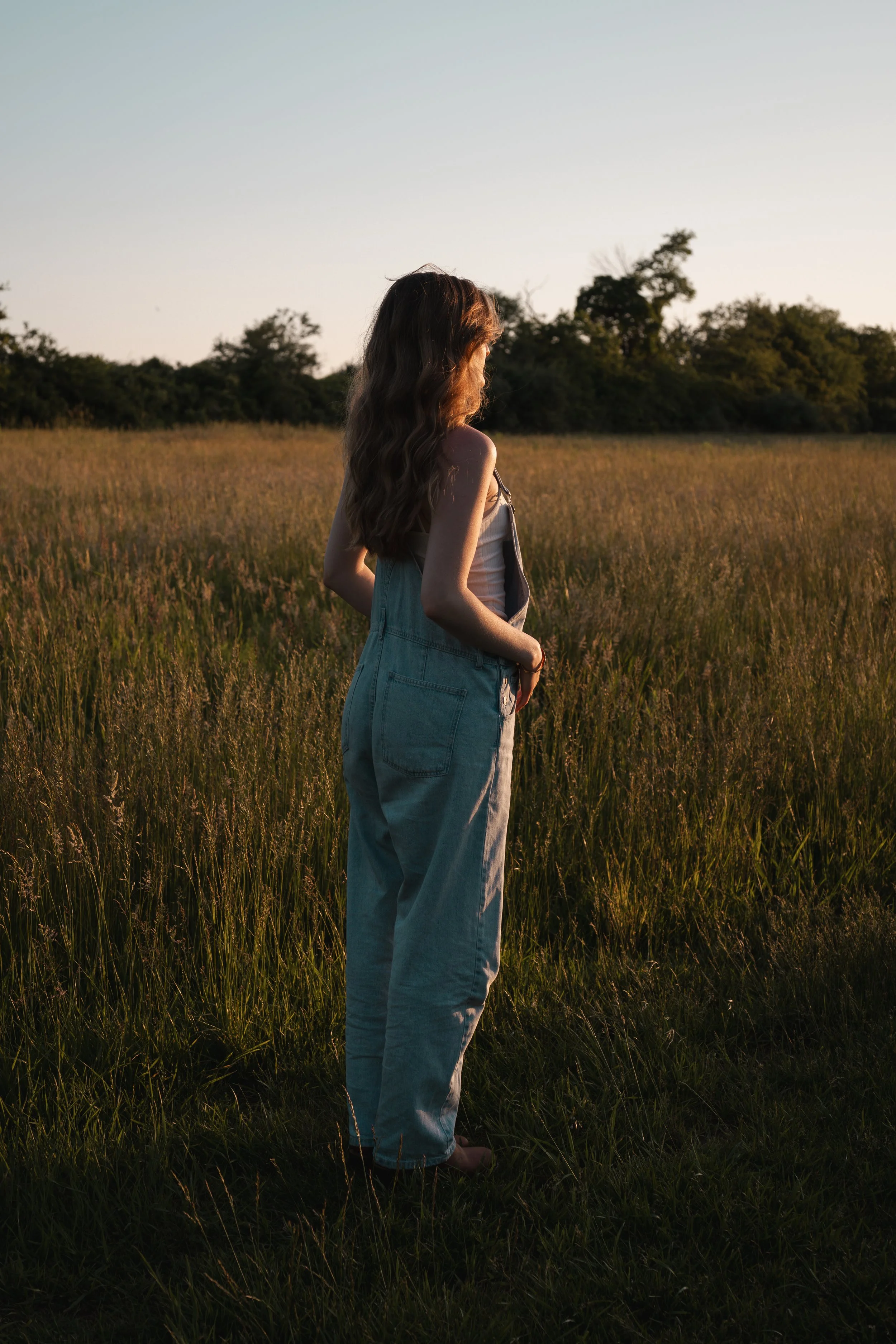 A woman with long brown hair, wearing a beige tank top and denim overalls, stands in a grassy field at sunset, looking to the side with her hands in her pockets.
