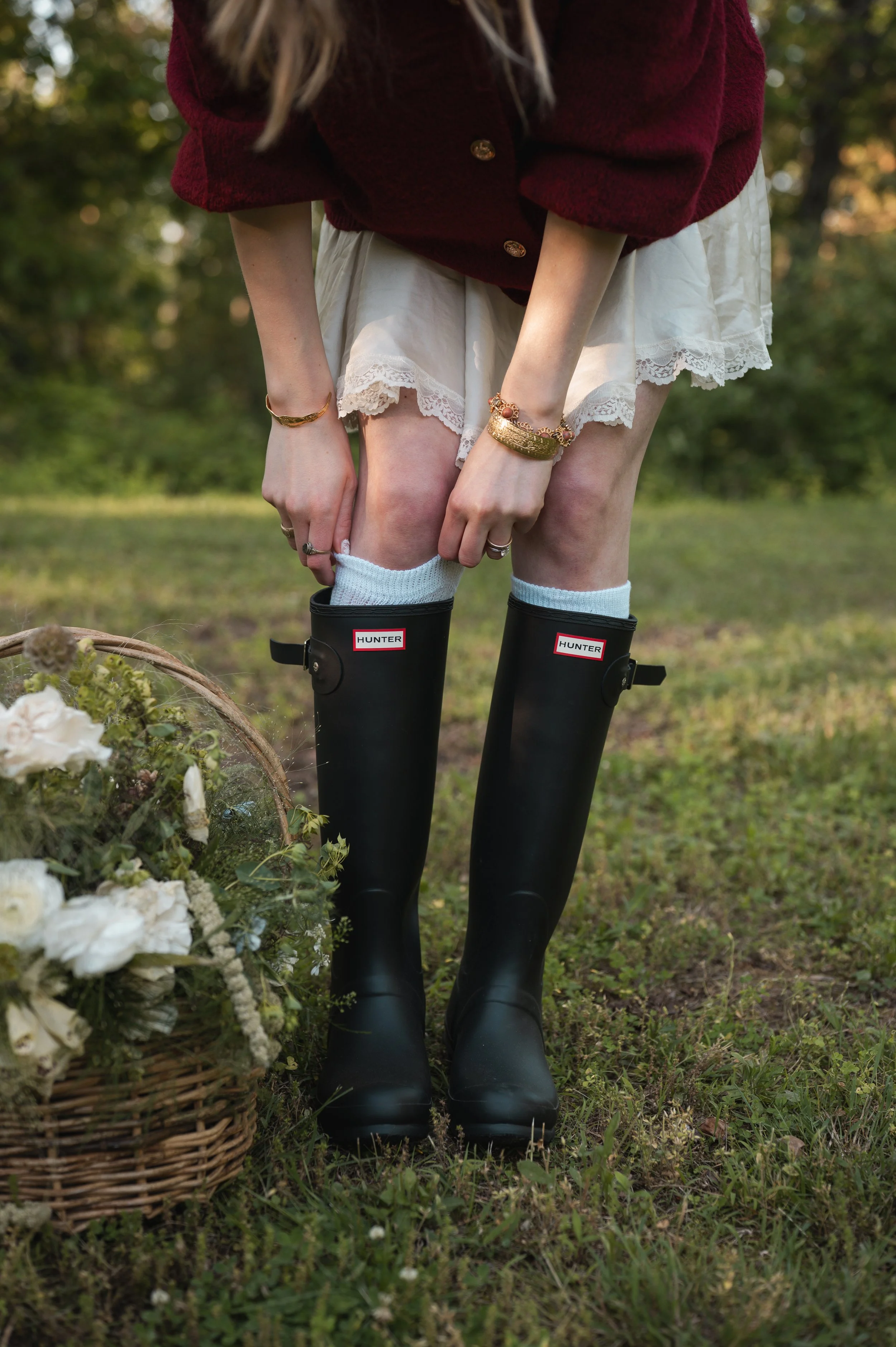 Person wearing black Hunter rain boots, white socks, a white lace dress, a burgundy sweater, and jewelry, standing outdoors on grass, near a basket of white flowers.