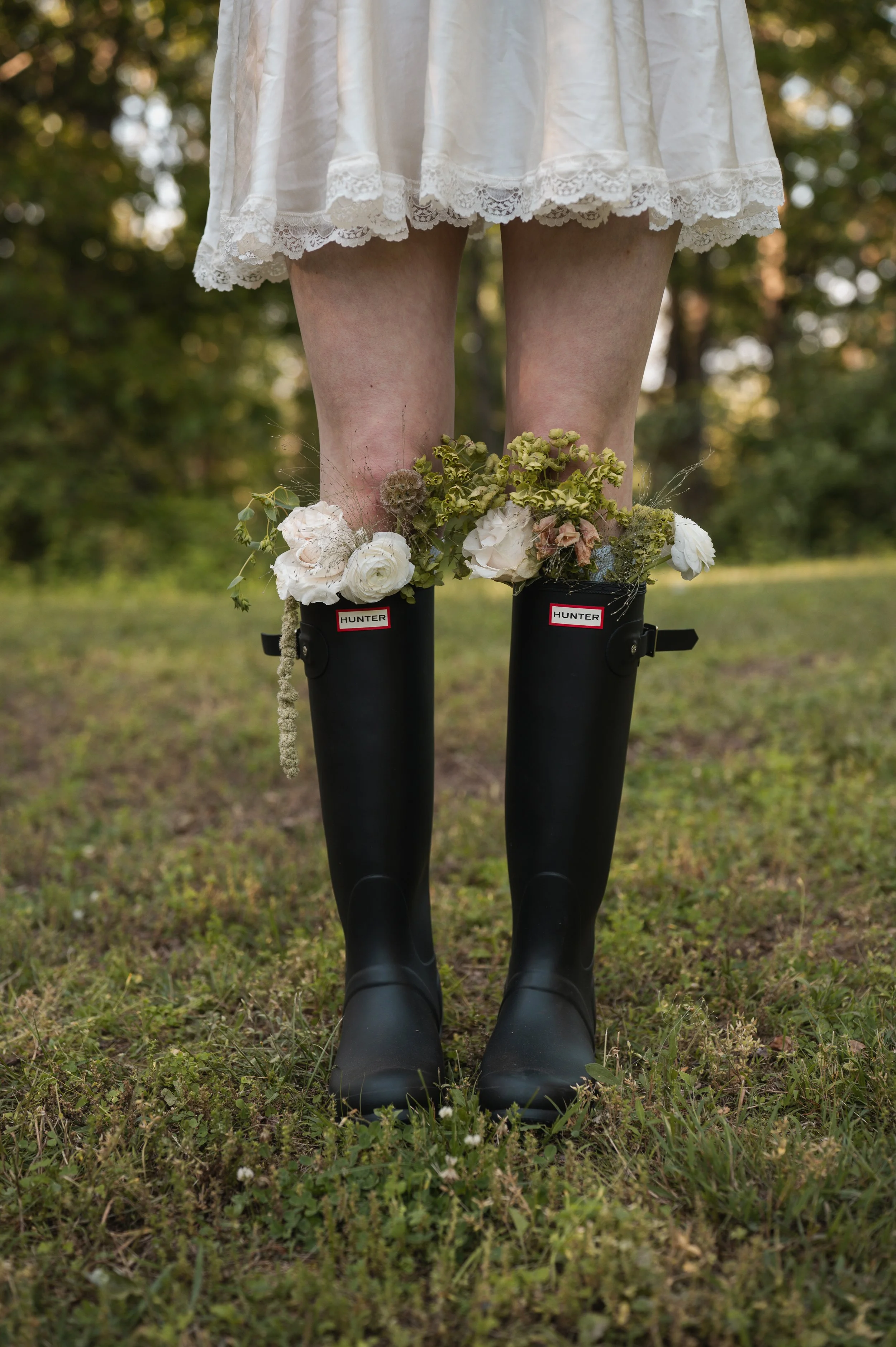 A person standing outdoors on grass, wearing a white lace dress, black Hunter rain boots decorated with flowers, and flowers around their ankles.