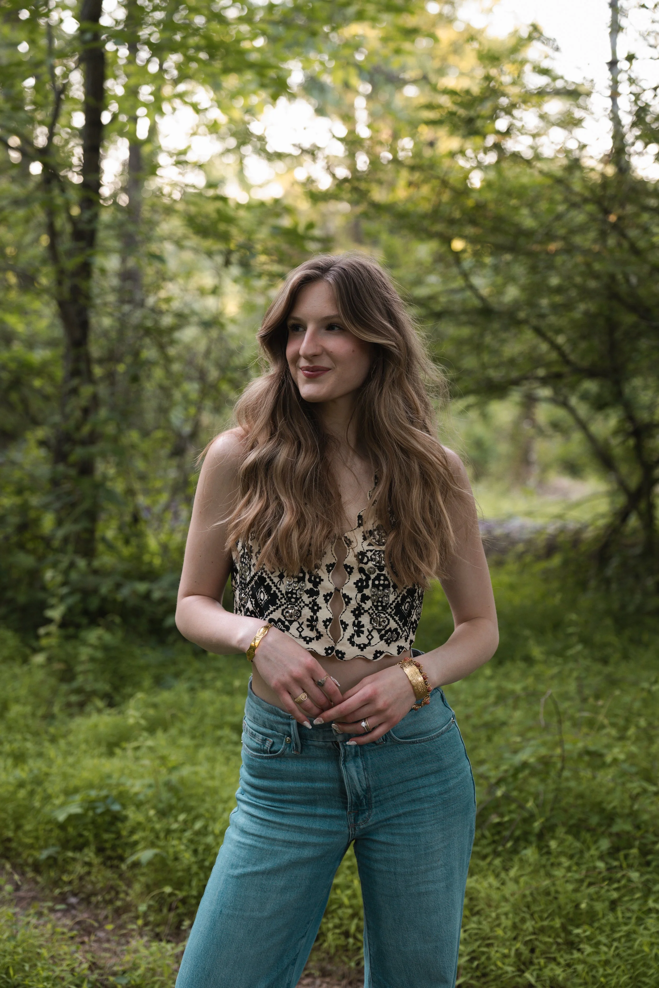 A young woman with long wavy brown hair standing outdoors in a forest with green trees and grass, wearing a sleeveless patterned top and blue jeans, smiling softly.