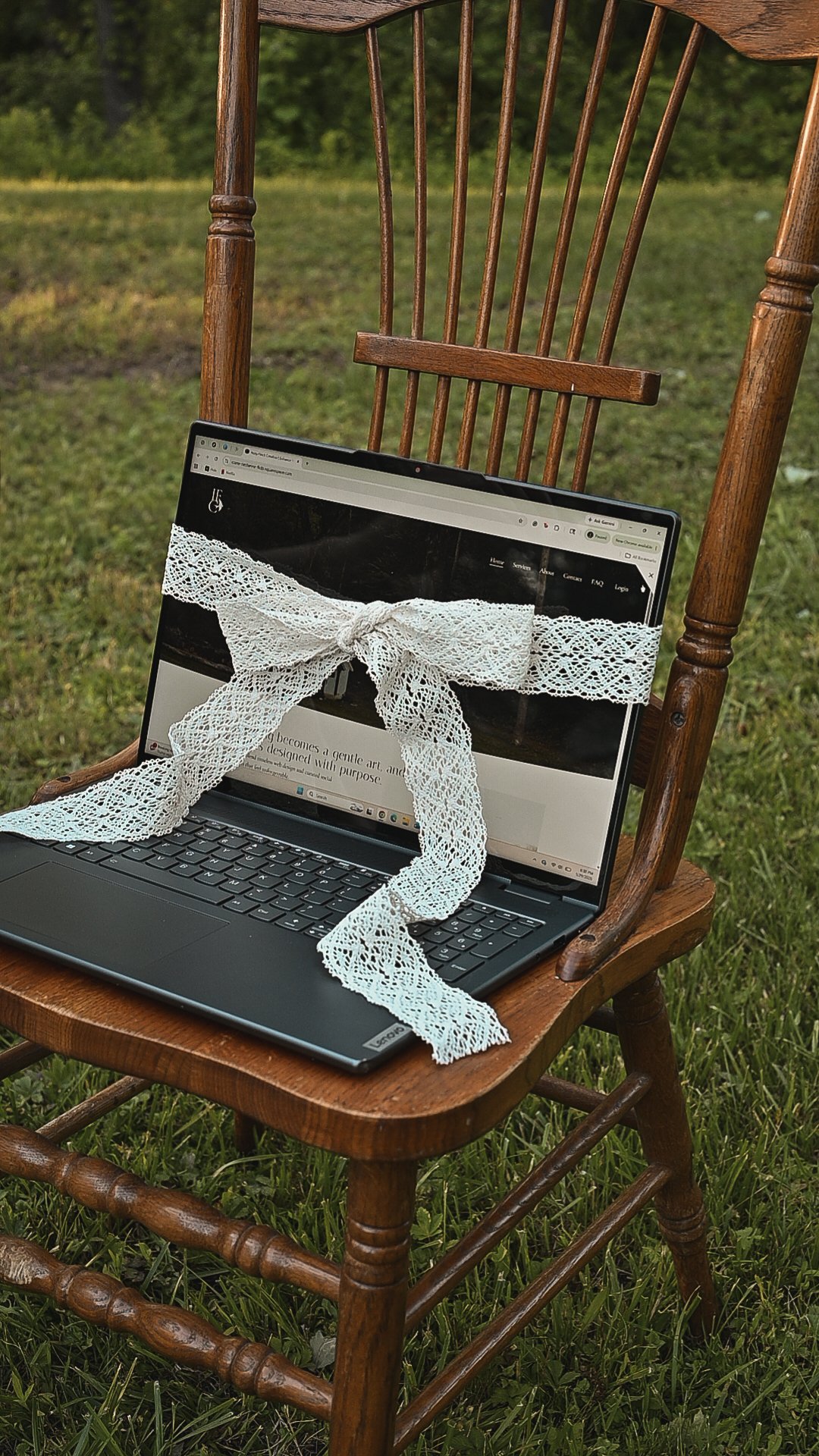 A laptop with a lace ribbon tied in a bow across the screen, placed on an old wooden chair outdoors on green grass.