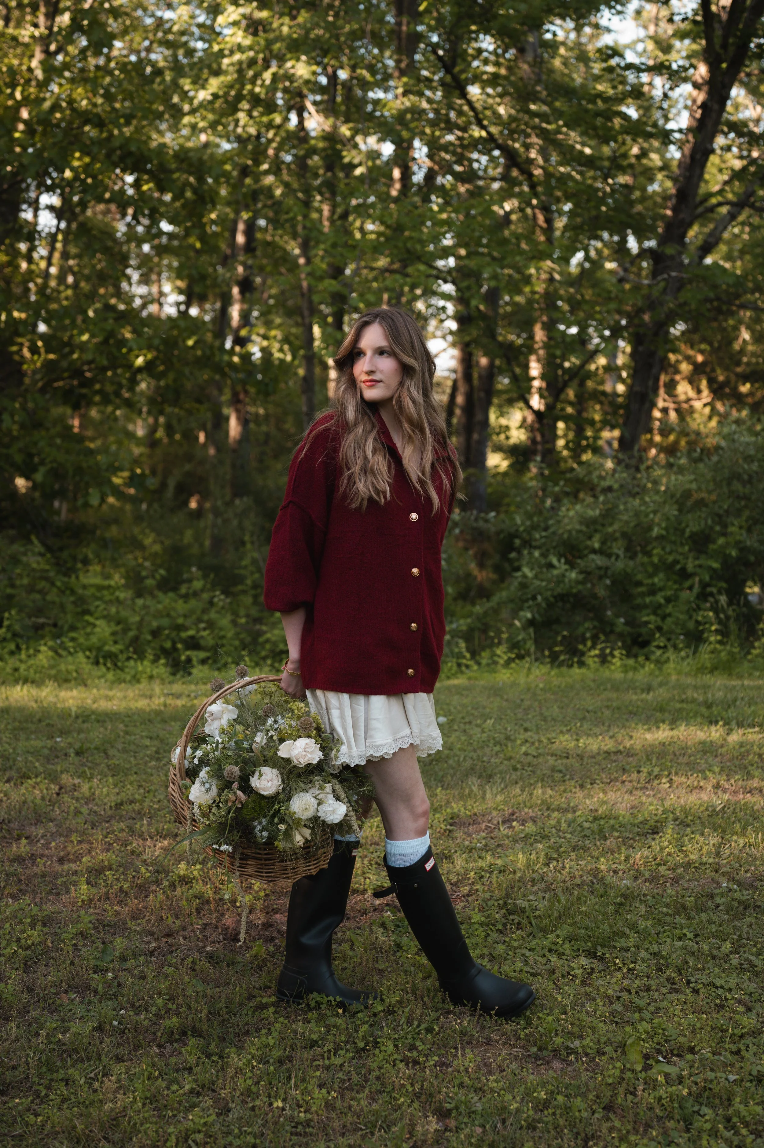Young woman in outdoor setting with trees, wearing a red jacket, white skirt, and black rain boots, holding a basket of flowers.