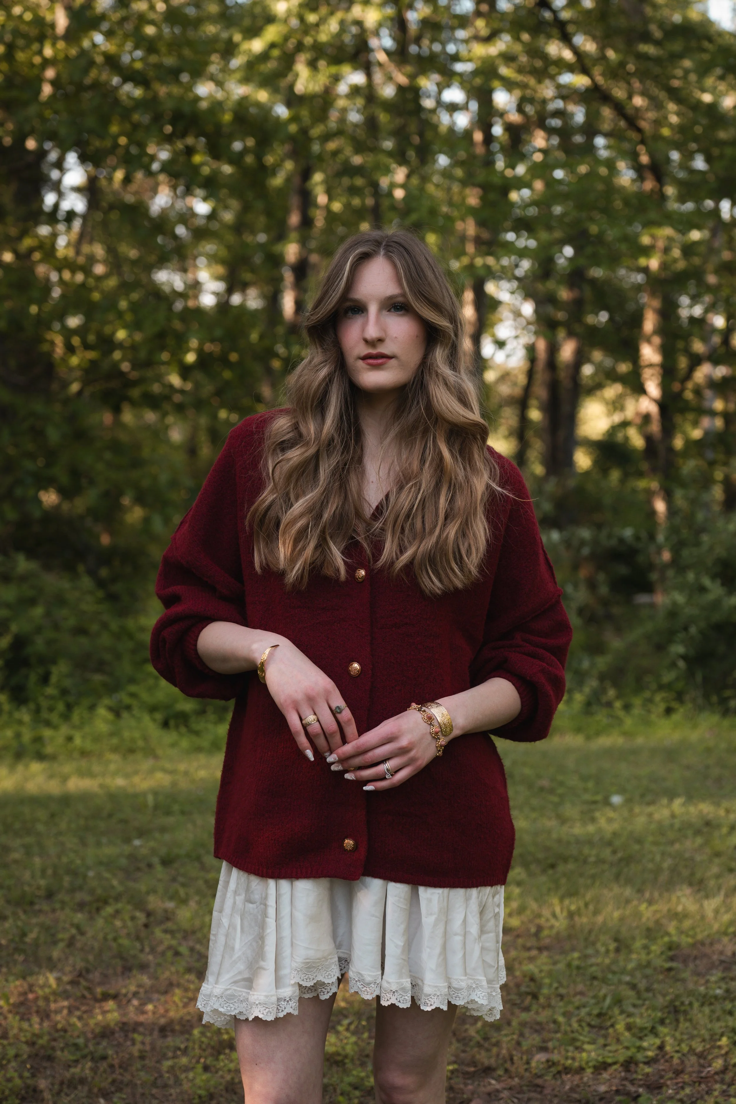 A young woman with long, wavy brown hair standing outdoors in a forested area with green trees in the background. She is wearing a deep red jacket, a cream-colored skirt with lace trim, and multiple gold jewelry pieces including rings, bracelets, and a watch. She has a neutral expression and is looking directly at the camera.